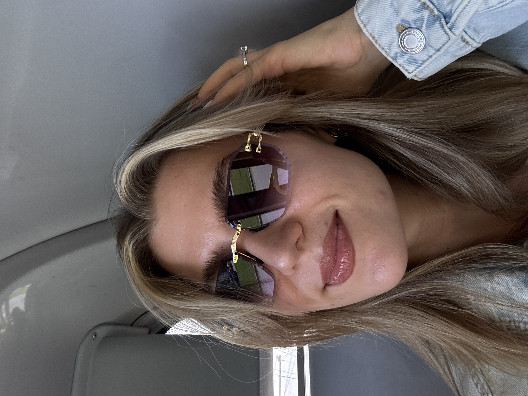 The image is a portrait of a woman, oriented on its side, showing her from the chest up. She has long, blonde and light brown hair, which falls around her face and over her shoulders. She is wearing large, frameless sunglasses with gold bridge and hinge details, which reflect an outdoor scene with what appears to be trees and possibly sky. Her lips are covered in a glossy, light pink or nude shade.
Her left hand is visible, resting in her hair near her forehead, and a silver ring with a single clear stone is visible on her ring finger. She is wearing a light blue denim jacket, the cuff of which is visible on the right side of the frame. A circular silver button on the jacket cuff has embossed text, partially discernible as "ORIGINAL" and "DENIM" or a brand name within a ring.
The background appears to be the grey interior of a vehicle, possibly a car, with smooth panels and subtle textures. The woman is looking directly at the camera with a slight smile, and her expression is calm. The lighting suggests it is daytime.