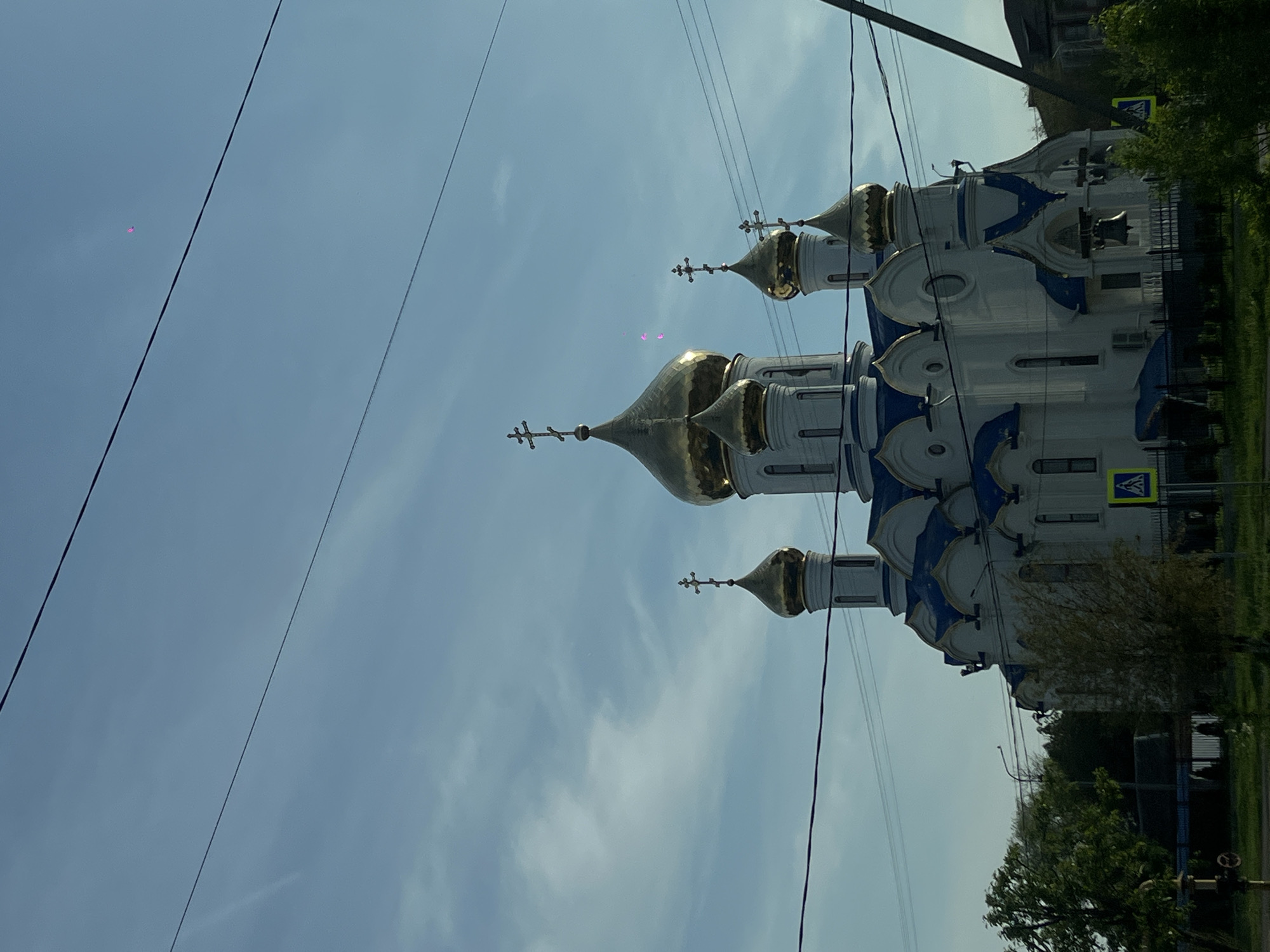 The image shows the Cathedral of the Intercession of the Most Holy Theotokos, located in Novopokrovskaya, Russia. The cathedral is white with blue accents and has golden domes with crosses. There are power lines in the foreground and trees and other buildings in the background. The sky is blue with some clouds.