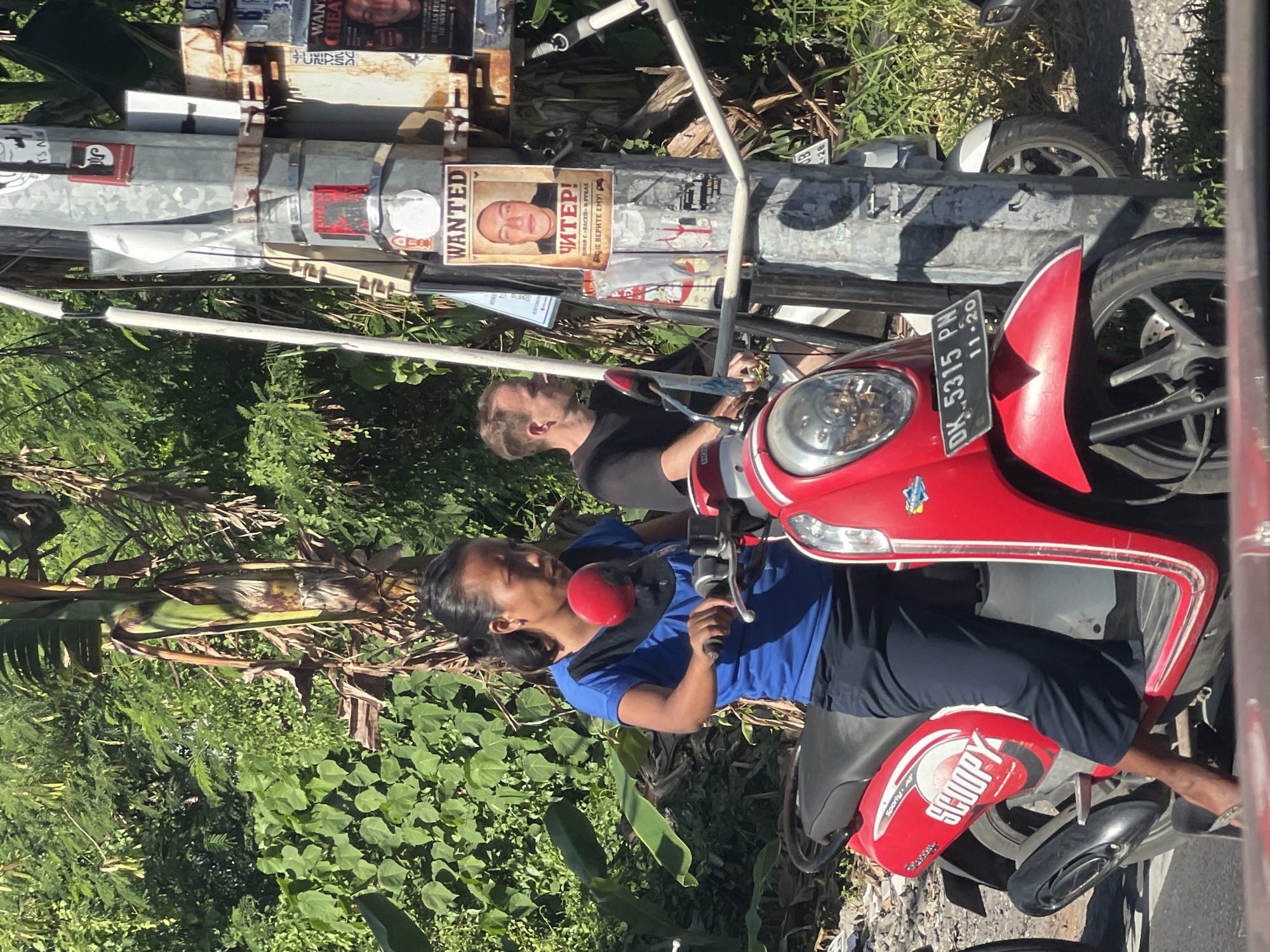 The image depicts two individuals on a red scooter in an outdoor environment, possibly in Unknown, Unknown, as suggested by posters and license plates present. The person in the foreground is operating the scooter and wearing a blue shirt and dark pants. In the background, another individual, wearing a black shirt, is seated behind the scooter operator. The scooter has a license plate that reads "DK. 5315 PN 11-20" and the word "SCOOPY" written on it. Adjacent to the scooter, there's a metal pole covered with various posters, including one that says "WANTED ЧИТЕР!" and another with the words "HE HILLS & LILLIES" and "HE NINTH". The surroundings include lush green vegetation and elements of an urban landscape.