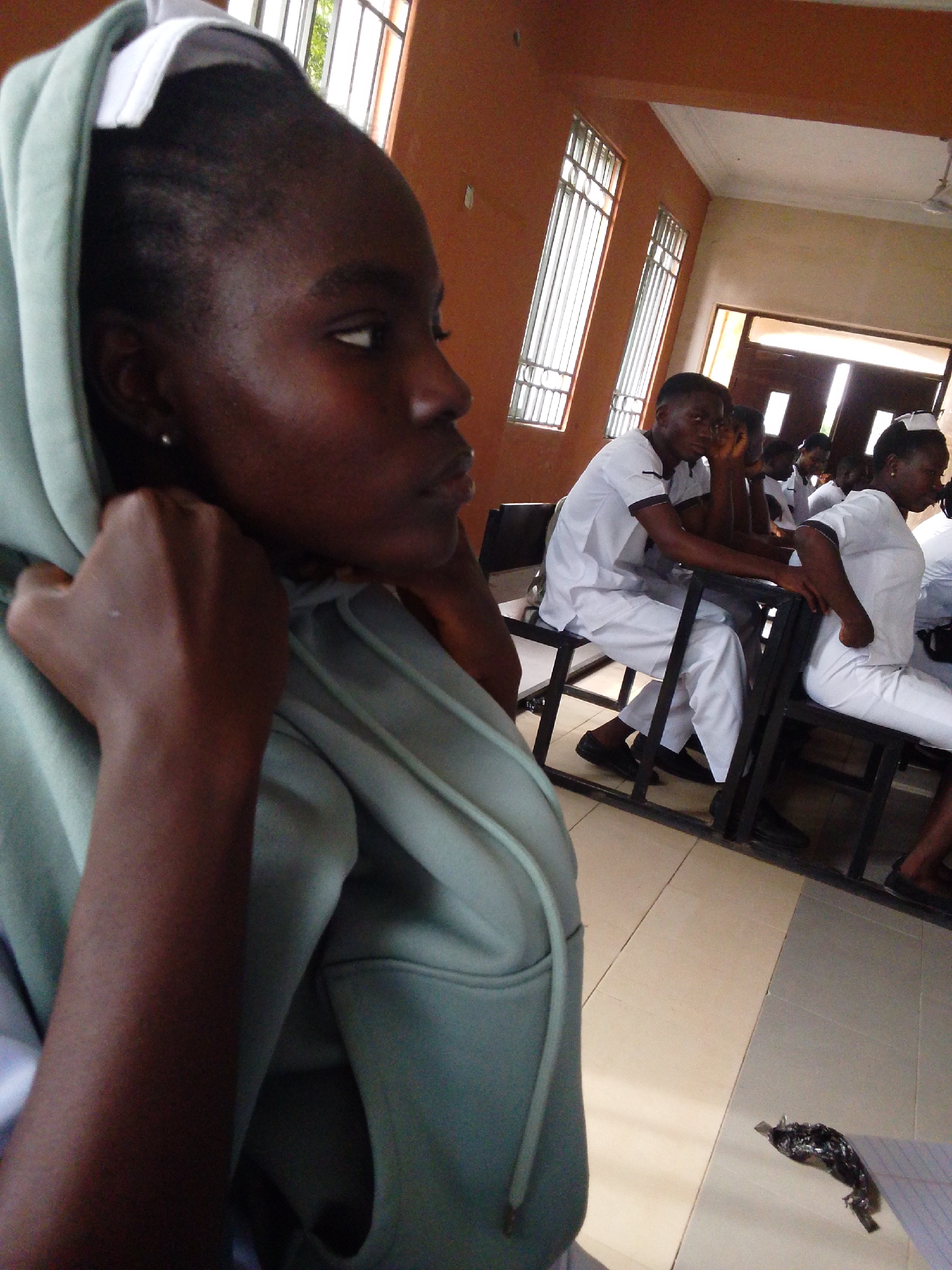In Kuson, Nigeria, within a classroom setting, a young woman in a light green hooded sweatshirt is the primary subject, positioned in the foreground. Her head is turned to her right, and she is adjusting the hood with both hands. She has dark skin, dark hair, and wears a small earring in her left ear. Behind her, several individuals are seated at desks, appearing to be students. They are dressed in white uniforms, suggesting a medical or nursing school. One young man in the mid-ground is looking to his left, resting his head on his hand. Another person, also in a white uniform, is seated to his right, facing away from the camera. The room features orange walls, tall windows with metal bars, and a tiled floor. A ceiling fan is visible in the upper right corner of the frame. On the floor near the foreground, a crumpled piece of foil-like material and a notebook are present. The lighting suggests an indoor environment, possibly during the daytime, with light streaming in from the windows.