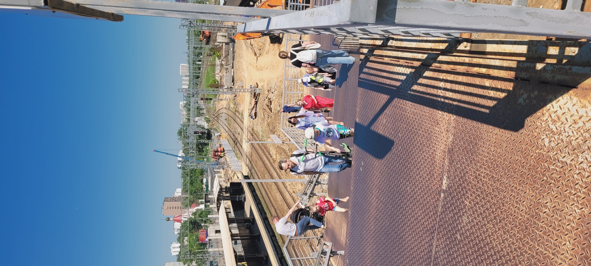 The image captures a sunny, elevated perspective of a pedestrian overpass overlooking a railway area in the city of Unknown, Unknown. In the foreground, a group of approximately ten individuals, including adults and children, are actively descending a set of rusted metal stairs with a diamond-plate pattern. On the far left, a man in a white t-shirt and jeans pushes a dark stroller while descending, followed by a barefoot child in a red shirt and blue shorts. In the center, an adult male in a light polo shirt and jeans holds a dark green kick scooter, while a child in a white t-shirt, patterned shorts, and a light green helmet also descends with a scooter. On the right, several people, including a woman in a light blue dress, a woman in a red dress, and a woman in a black t-shirt and jeans with a white backpack, are also descending. Sunlight from the left casts long, distinct shadows of the handrails and people onto the stairs.
Beyond the stairs, the midground reveals a complex railway infrastructure. Multiple tracks run parallel, flanked by train platforms with shelters on the left. Extensive overhead gantry structures supporting railway electrification are visible, along with several red signal lights. To the right of the tracks, a large area of disturbed earth indicates ongoing construction or renovation, with piles of dirt, construction materials, and heavy machinery, including an orange excavator, present. A tall blue crane is visible in the distance over the railway lines. The background features various urban buildings, including residential structures and a distinctive church with red domes and a spire on the far left. The sky is clear and bright blue.