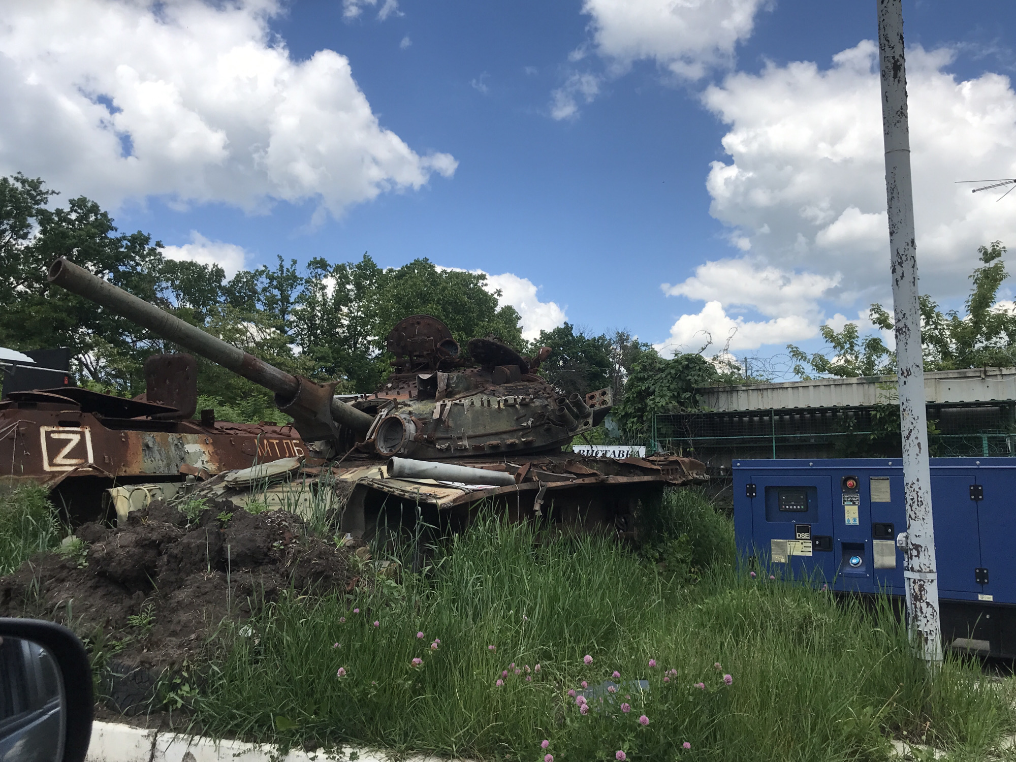 The image captures an outdoor scene featuring two heavily damaged and rusted military vehicles under a bright blue sky with scattered white clouds. In the foreground, a derelict tank or armored vehicle is partially overgrown with green grass and small pink clover flowers, and sits atop a mound of dark earth. Its main gun barrel is visible, pointed slightly upwards. The vehicle's turret appears heavily damaged or dislodged. Visible on its side are the Cyrillic letters "МТЛБ" (MTLB). Behind this vehicle, another equally rusted armored vehicle is partially visible on the left, bearing a white "Z" symbol within a square.
The setting appears to be a display or collection of destroyed war equipment. A partially visible sign behind the foreground vehicle shows Cyrillic text, with "ВИСТАВКА" (VYSTAVKA), meaning "EXHIBITION," being discernible. To the right, a tall, white utility pole with peeling paint stands prominently. Adjacent to the pole is a large, blue industrial generator with "DSE" visible on its panel. In the background, lush green trees frame the scene, and a fence topped with barbed wire runs along a concrete structure. The time of day is daytime, with clear, sunny weather. There are no people visible in the frame. The scene evokes a somber reflection on conflict and its aftermath.