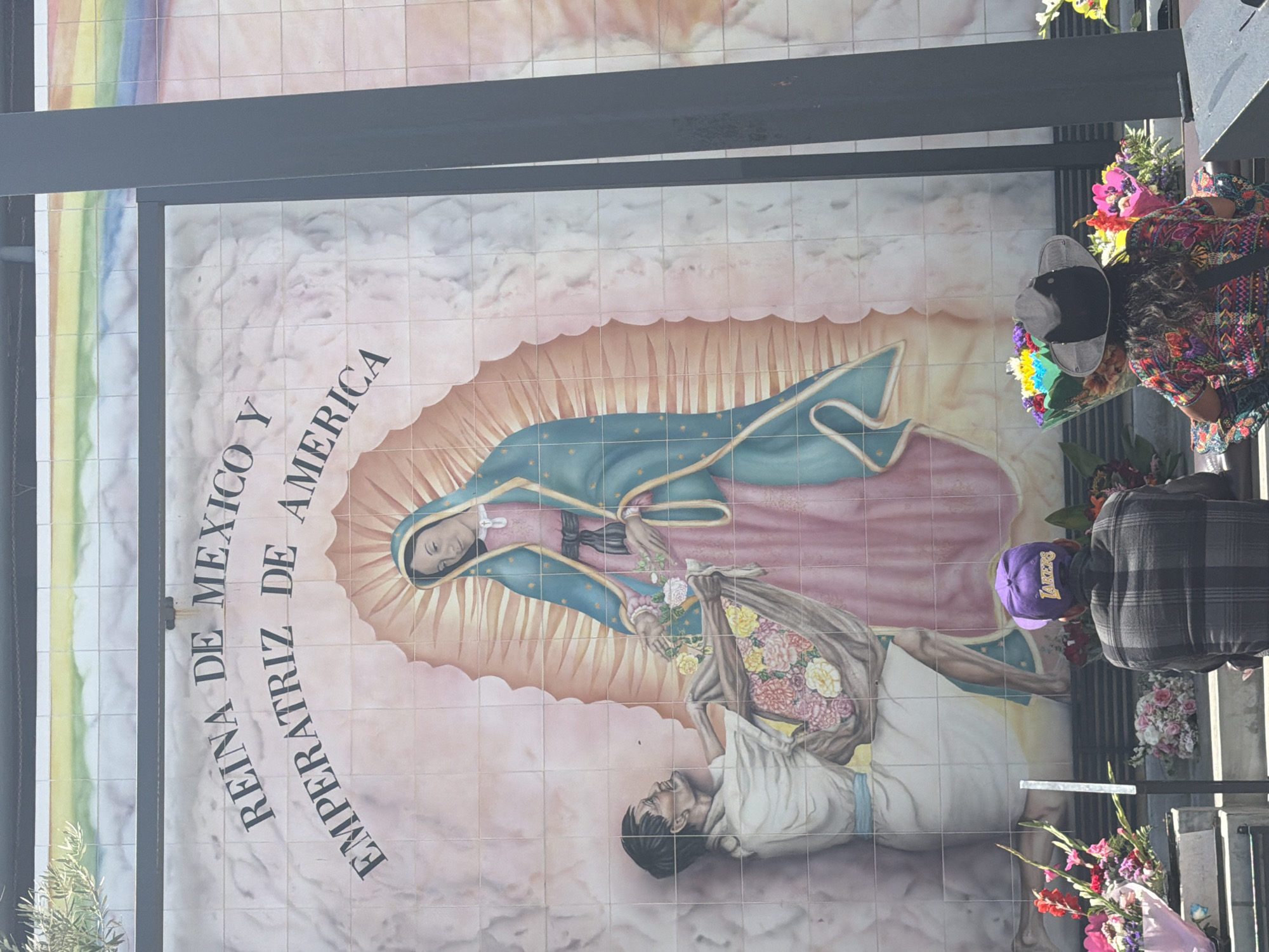 A mural depicting the Virgin of Guadalupe with Juan Diego as a tile mosaic. The text in Spanish translates to "Queen of Mexico and Empress of America". It is located in Los Angeles, California, indicated by the presence of a person wearing a Lakers hat and the diverse cultural elements. Mourners stand nearby with flowers.