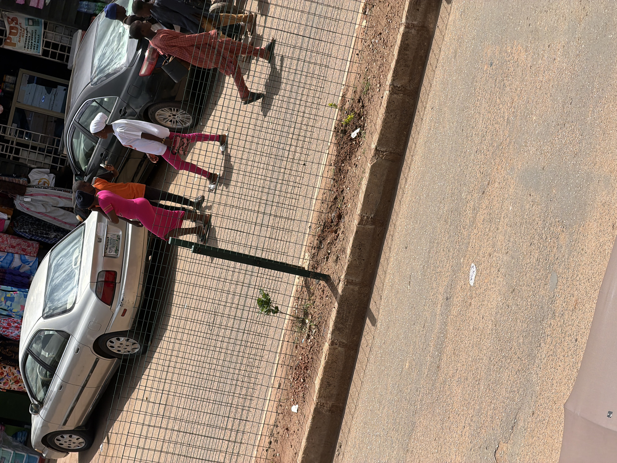 The image captures an outdoor scene in Bukuru, Nigeria, likely on a sunny day. A group of people are visible, some walking and others standing near vehicles. In the foreground, a silver sedan is parked on a paved surface, partially obscured by a wire fence. Behind it, a dark-colored car is also parked. Several individuals are walking along the paved area behind the fence. One person is wearing a red and white checkered shirt and dark pants. Another is dressed in a white top and pink pants, with a white headscarf. A third individual is wearing a bright pink dress. Further back, more people are gathered. The setting appears to be a street or market area, with buildings and shops visible in the background. The ground is a mix of paved surfaces and dirt. The sunlight casts distinct shadows, indicating it is daytime. No specific emotions are discernible from the individuals' postures or expressions. There is some visible text on a sign on one of the buildings in the background, but it is too blurry to read. The license plate on the silver sedan is partially visible and appears to have characters like "S-0704".