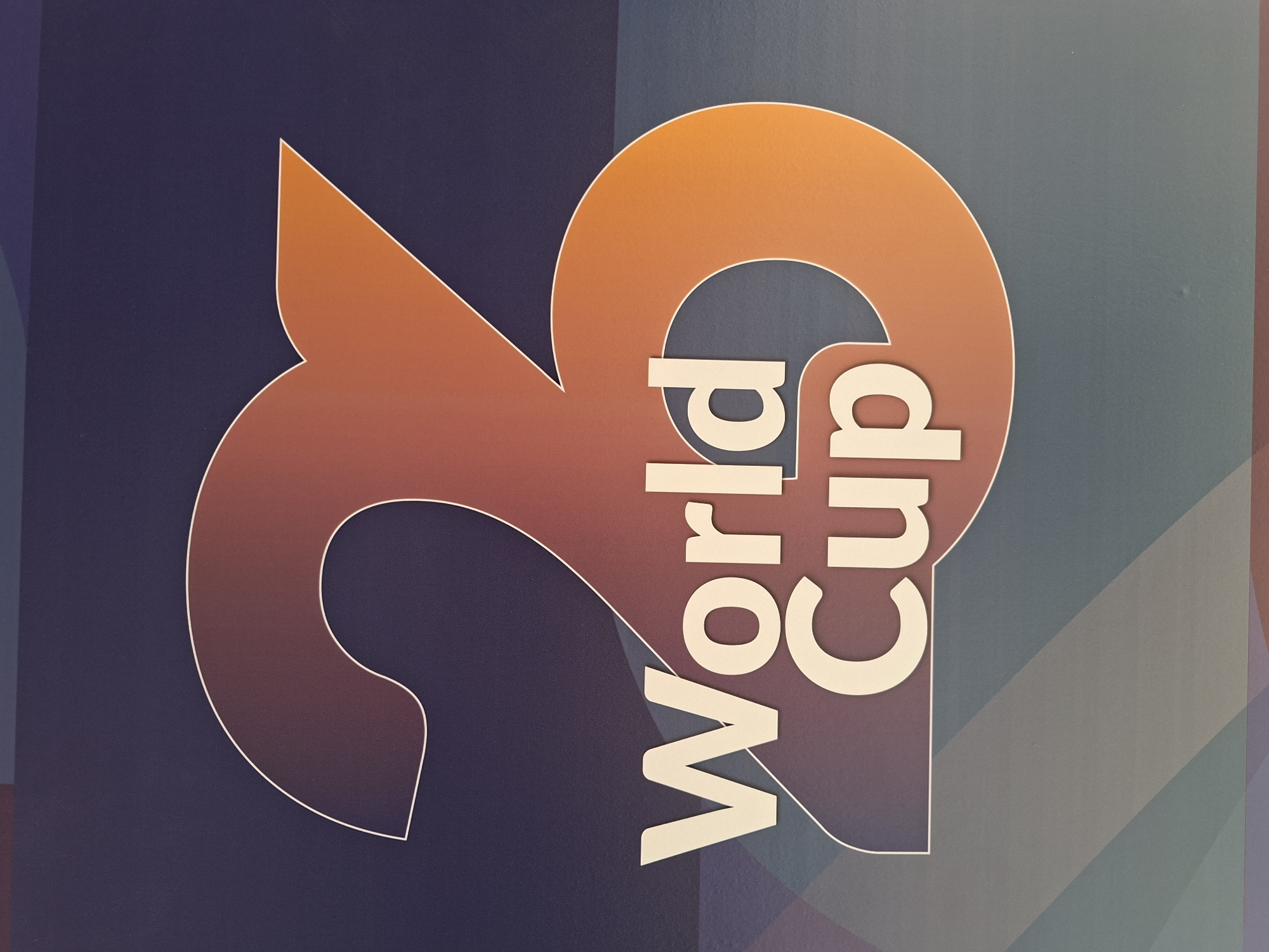 The image is a close-up, medium shot of a logo that reads "23 World Cup". The logo is large and features gradient colors ranging from purple to orange. The numbers "23" are prominent and stylized, with the "2" appearing on the left and the "3" on the right. Below the numbers, the words "World Cup" are written in white, sans-serif font. The background is a dark, textured surface with subtle diagonal lines of muted colors. The logo appears to be printed or displayed on a wall or banner. The exact location in Al Rayyan, Qatar is not discernible from the image itself. There are no people or actions depicted.