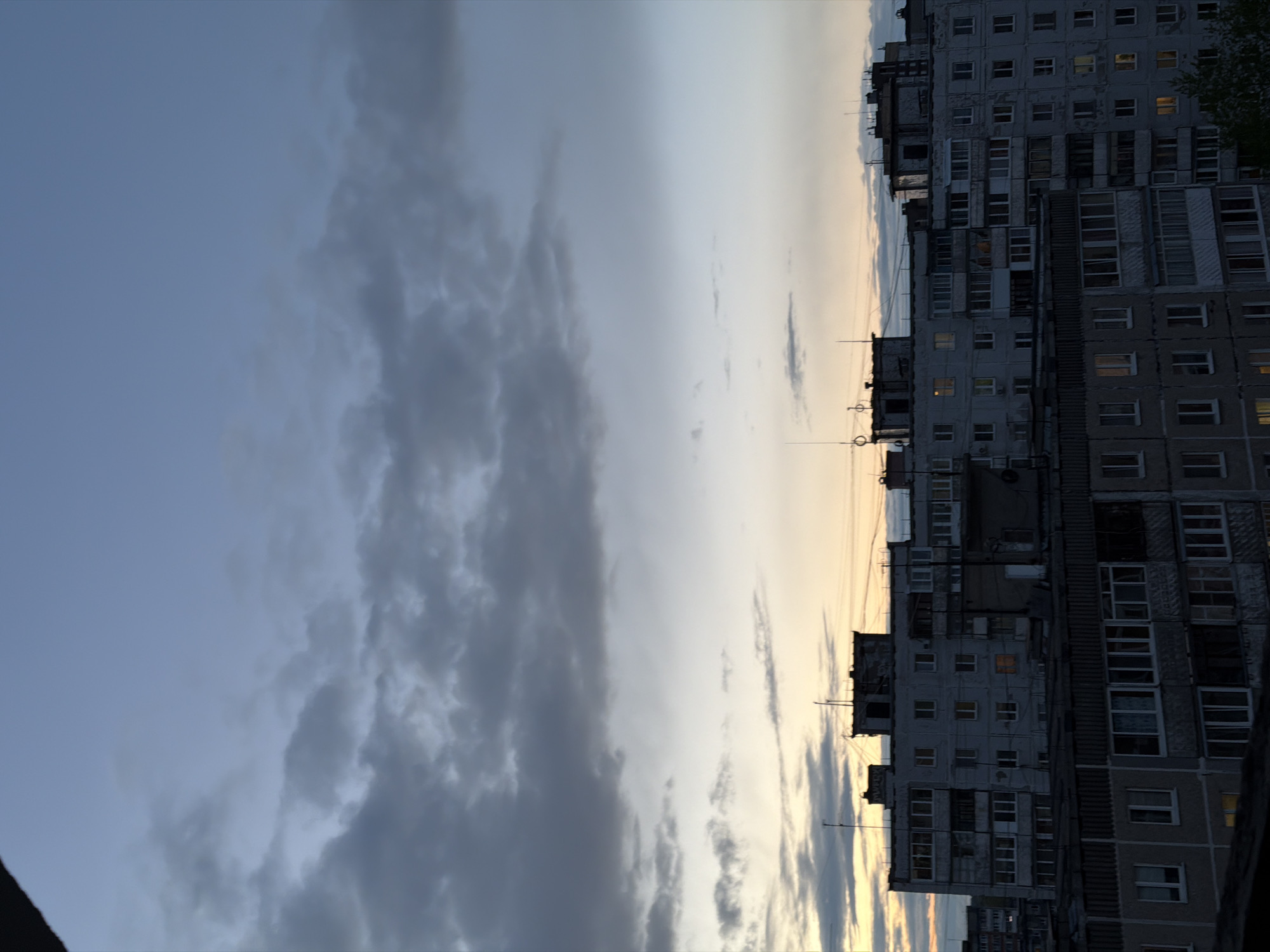 The image depicts a high-rise apartment building in Perm, Russia, as seen from an elevated perspective. The building is tall, with numerous windows arranged in a grid-like pattern, some of which are illuminated. The building's facade is gray and appears weathered. The sky is overcast, with dark, heavy clouds dominating the left side of the frame and a faint golden hue indicating either sunrise or sunset on the right. Antennas are visible on the building's roof. There are no people visible in the shot. The environment is urban.