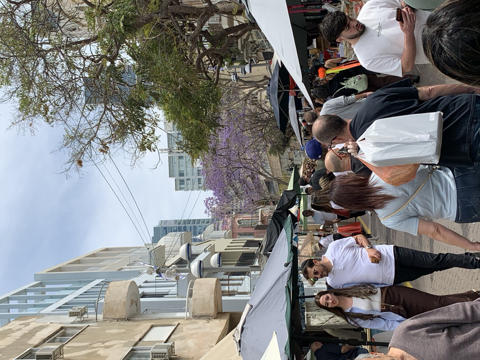 This image captures a vibrant daytime scene at an outdoor market or street fair in Tel-Aviv, Israel. The street is bustling with numerous people, suggesting a lively event.
In the foreground, a man with a beard, wearing sunglasses and a kippah, stands in a white t-shirt and dark pants, looking to the left. Beside him, a woman with long dark hair, dressed in a light blue shirt over a white top and brown pants, smiles gently. Further into the crowd, a man in a black t-shirt and dark cap is seen carrying a large white plastic bag containing a rectangular item, along with an orange plastic bag. Another man in a white t-shirt is observed looking at his mobile phone. Many other individuals are visible, either walking, browsing, or partially obscured by the crowd and market stalls. Most people are dressed casually.
The setting includes a row of market stalls with white and dark awnings, under which people are congregating. The street is lined with a variety of buildings, ranging from older, multi-story structures featuring balconies and visible air conditioning units, to modern high-rises in the background. Overhead, utility wires crisscross the clear, bright sky. Several trees are prominent, including one with distinctive vibrant purple blossoms, likely a Jacaranda, adding a striking color accent to the urban landscape. Other trees have green foliage or bare branches.
The activity taking place is typical of a street market or fair, with people engaging in shopping, strolling, and socializing. The bright, even lighting indicates it is daytime with pleasant weather. While some faint text is visible on a plastic bag and a man's t-shirt, it is not clearly decipherable. The overall mood appears relaxed and active.