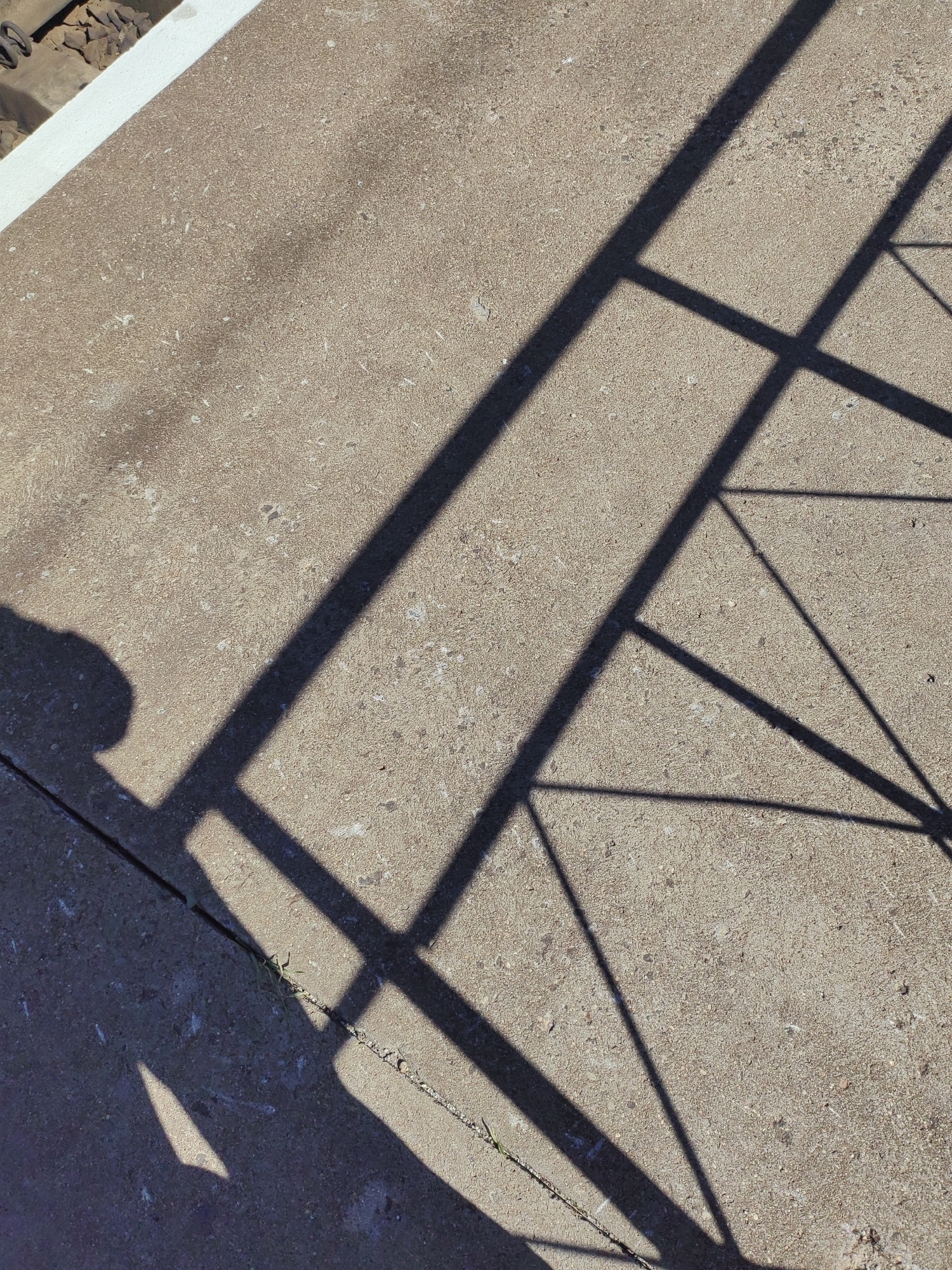 This media file captures a close-up view of a concrete surface, likely a pathway or sidewalk, with distinct shadows cast upon it. The shadows form geometric patterns, suggesting the presence of a railing or fence. One prominent shadow appears to be of a person's head and shoulder, casting a silhouette onto the ground, indicating a person is present and standing or walking in the scene. The overall scene is outdoors, under bright sunlight, as evidenced by the sharp and defined shadows. The time of day appears to be mid-morning or late afternoon, judging by the angle of the shadows. The concrete itself shows signs of wear and tear, with small stones and imperfections visible, typical of an outdoor public space. In the upper left corner, a glimpse of railway tracks can be seen, partially obscured by a white edge. This suggests the location might be near a railway line, possibly a platform or a crossing. The overall mood conveyed by the image is one of simple, everyday observation, focusing on the interplay of light and shadow on a mundane surface. There is no visible text in the image.