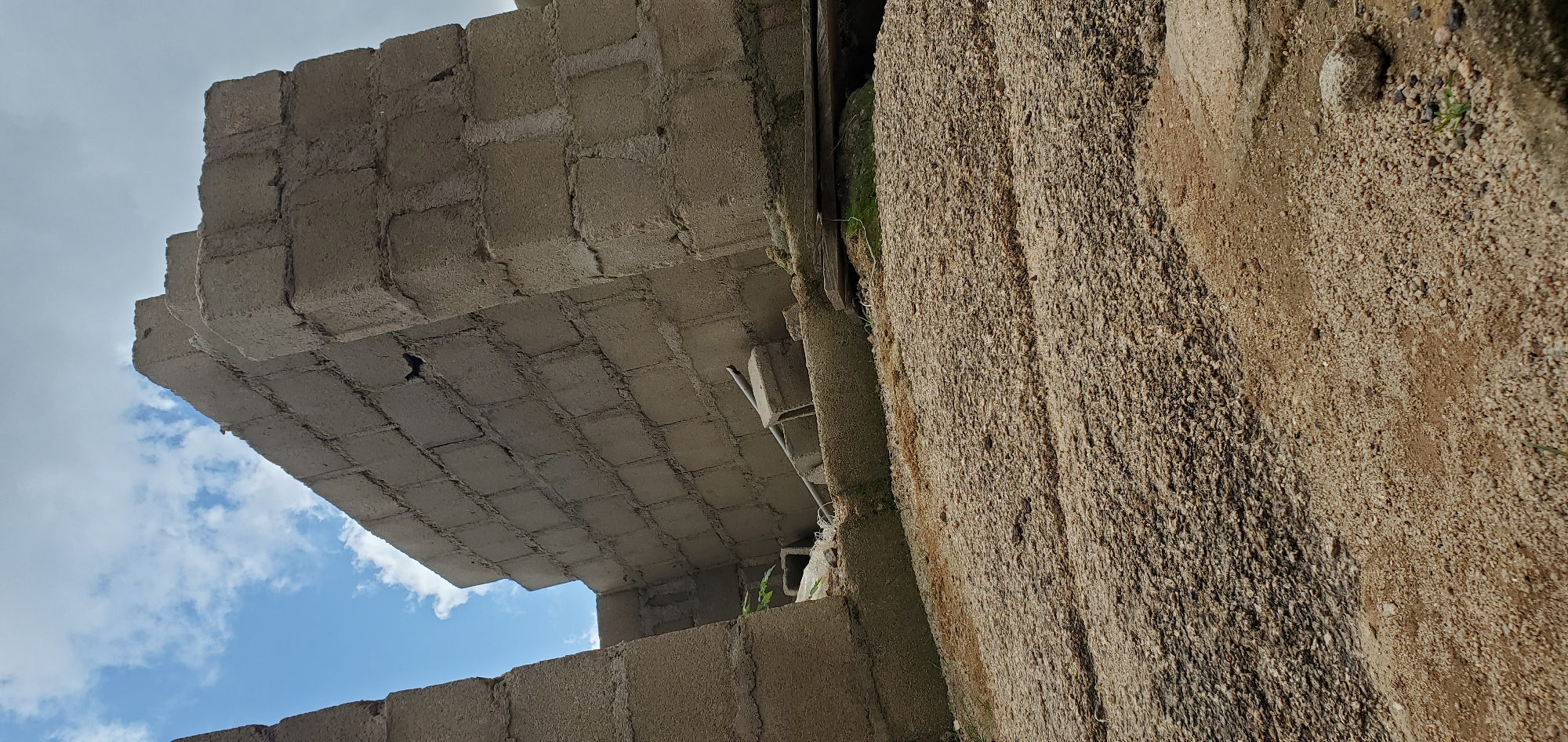 The image depicts the partially constructed and leaning walls of a building, made from concrete blocks, under a partly cloudy sky. The foreground is covered with gravel and sand. There are no people visible in the image. The scene appears to be outdoors in an undeveloped or construction area.