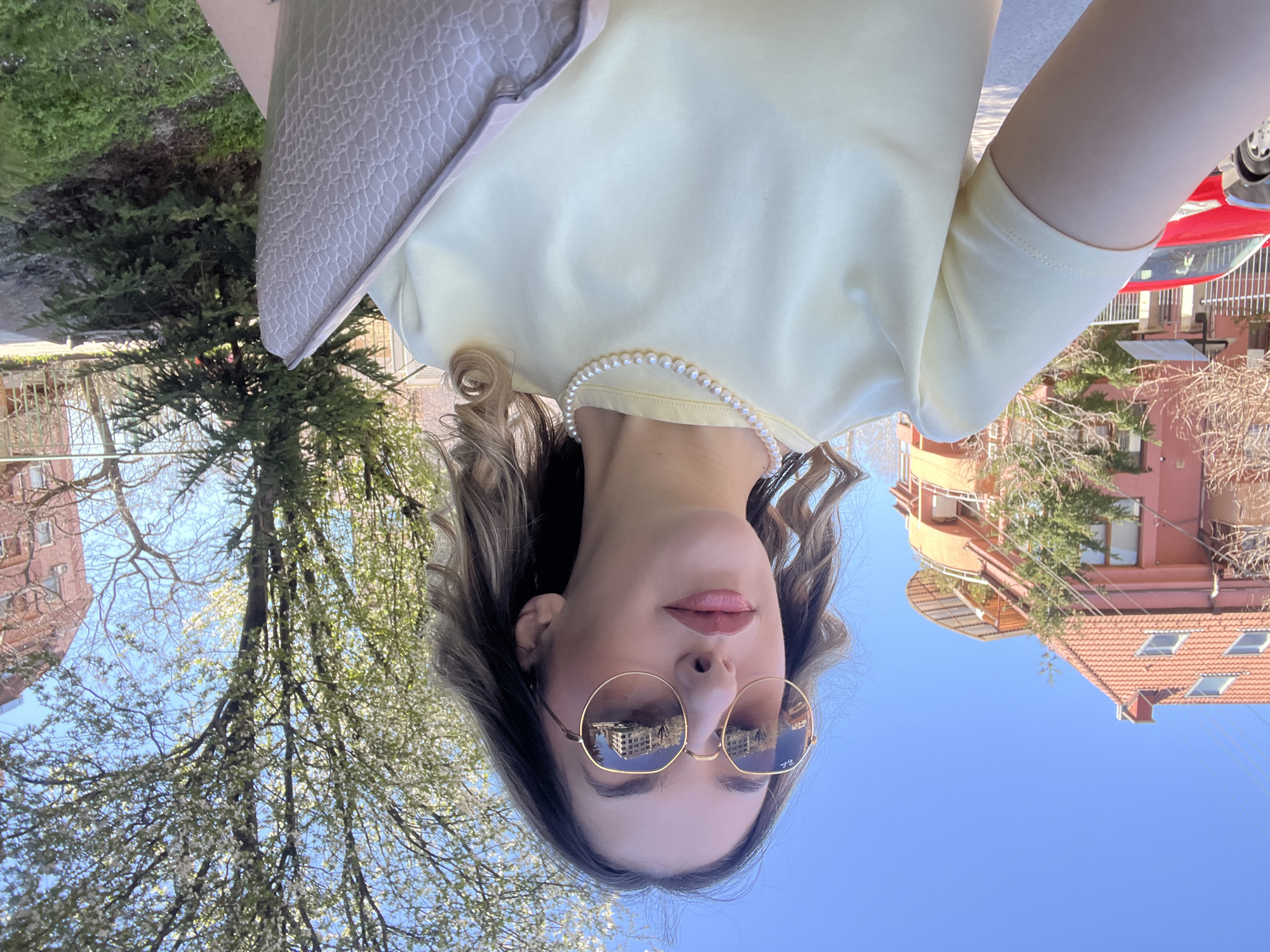 A woman is centered in the frame, wearing a light yellow top and a pearl necklace. She is also wearing round-framed sunglasses that reflect buildings. Part of a textured, light gray purse is visible at the top left of the frame. Behind her are trees, buildings, and blue sky. The buildings reflected in her sunglasses appear to be located in Sofia, Bulgaria, based on their architectural style. A red car is visible in the top right corner.
