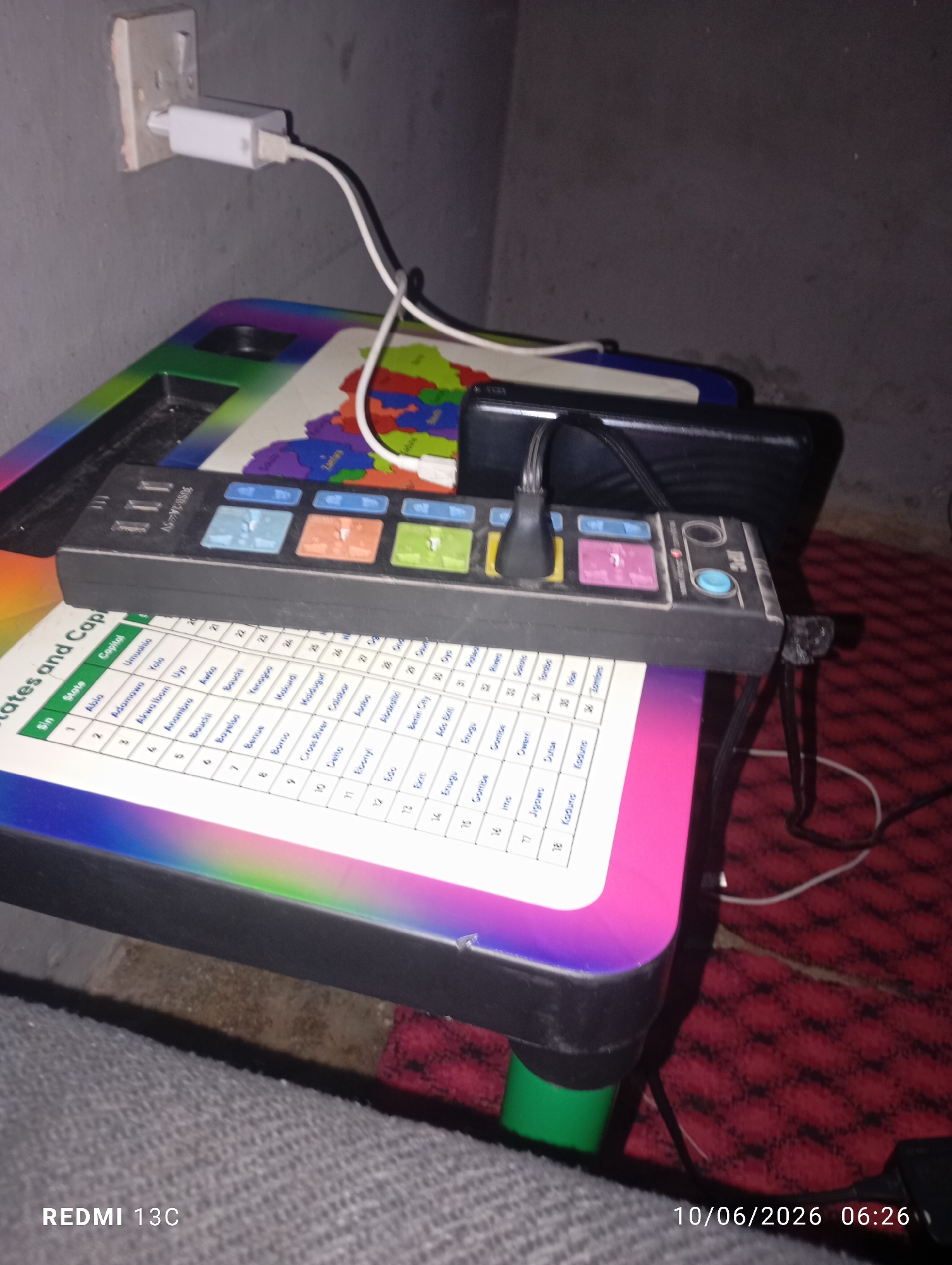 A close-up shot captures a colorful, foldable desk with a map of Nigeria on its surface. On the desk rests a black power strip with multiple outlets and colorful buttons, one of which is illuminated. A white USB charging cable is plugged into the power strip and extends upwards to a white power adapter connected to a wall socket. A black smartphone or electronic device lies next to the power strip. The desk is placed on a red textured rug. The background wall appears to be rough and gray. The timestamp "10/06/2026 06:26" and the text "REDMI 13C" are visible in the bottom corners of the image. The scene is likely indoors, possibly in a residential setting. The presence of the map of Nigeria and the timestamp without specific location context in the image itself means it is not possible to determine the location in Jos, Nigeria.