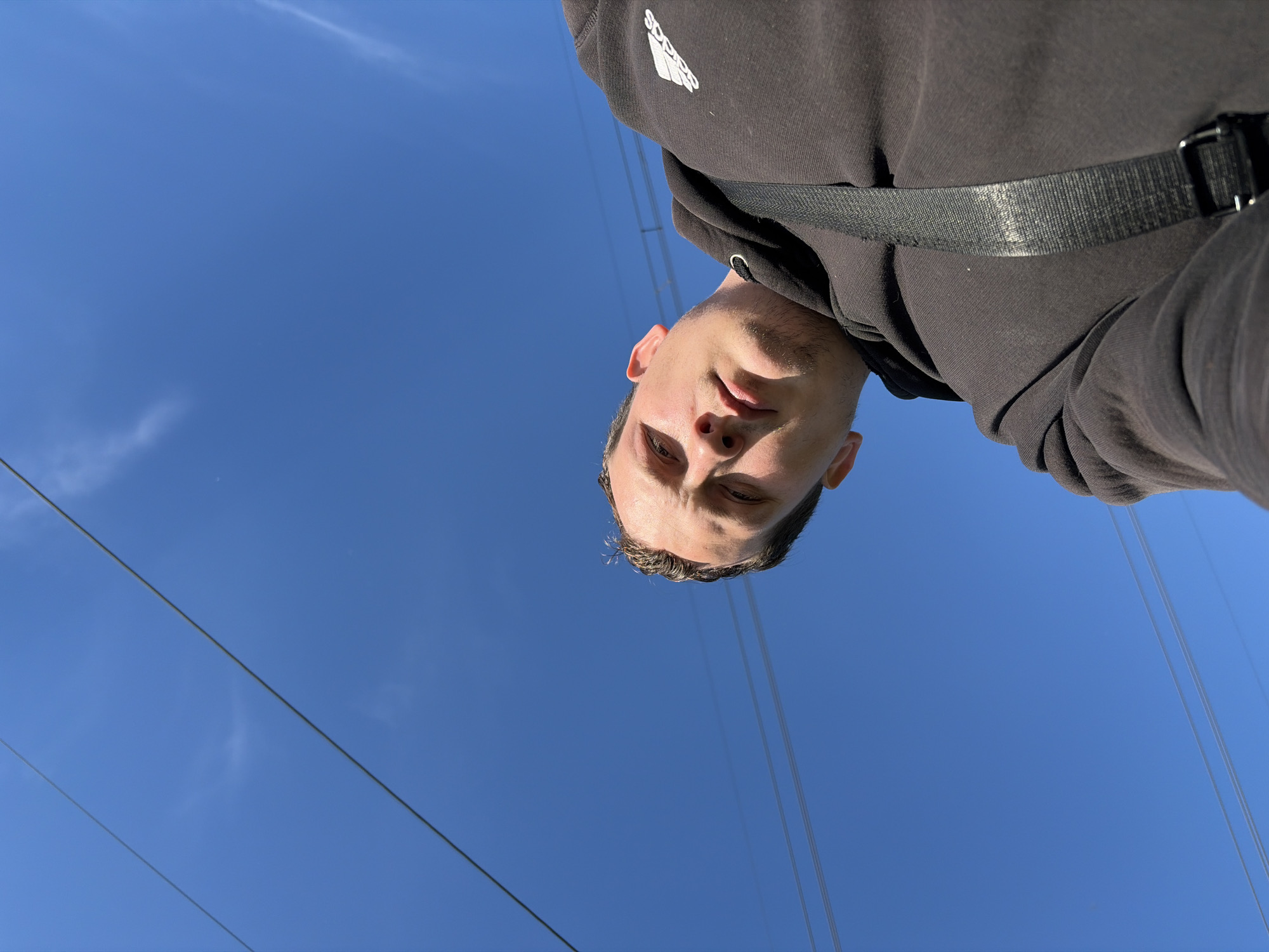 A low-angle, upward-facing shot captures a man, likely taking a selfie, against a bright blue sky. The man is wearing a dark-colored hoodie with the Adidas logo visible on the upper left portion of the fabric. A black strap crosses his torso. Power lines stretch across the sky. The location is Chaikovsky, Russia.