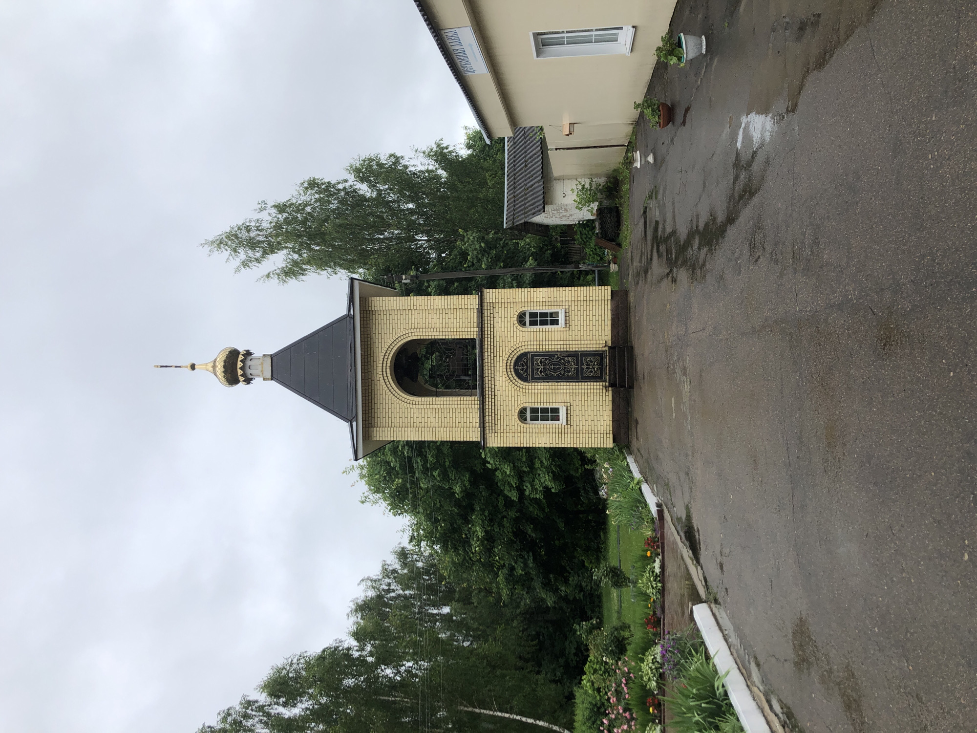 The image shows a bell tower in Safonovo, Russia, on a cloudy day. The bell tower is made of yellow brick and has a black roof with a golden dome. In front of the bell tower, there is a paved walkway with some puddles, indicating recent rain. To the left of the walkway, there is a flower bed with various colorful flowers. To the right of the bell tower, there is a long, light-yellow building with a sign that reads "ЦЕРКОВНАЯ ЛАВКА" (Church Shop). Behind the bell tower and the shop, there are several trees and a smaller white building. There are no people visible in the image.