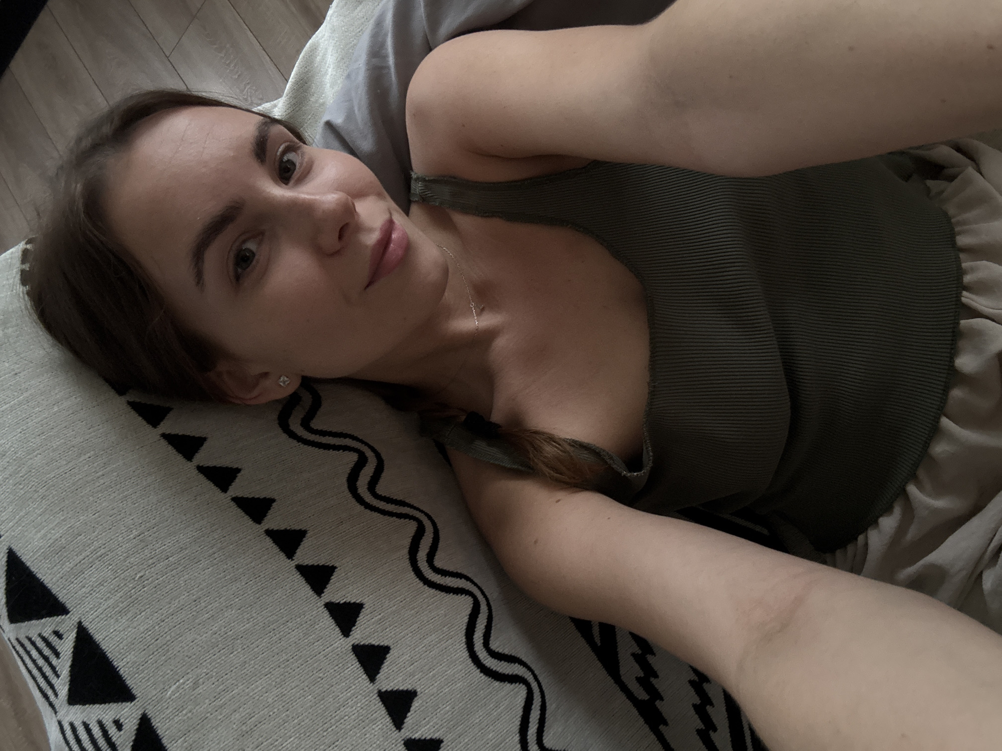 A young woman with light brown hair tied in a braid and wearing a green ribbed tank top and light-colored shorts is lying on a bed. She is looking at the camera and smiling. The bed has a gray pillow and a blanket with a black and white geometric pattern. The floor appears to be made of light-colored wood. The image is taken from a high angle, looking down at the woman. The scene is likely indoors, in a bedroom.