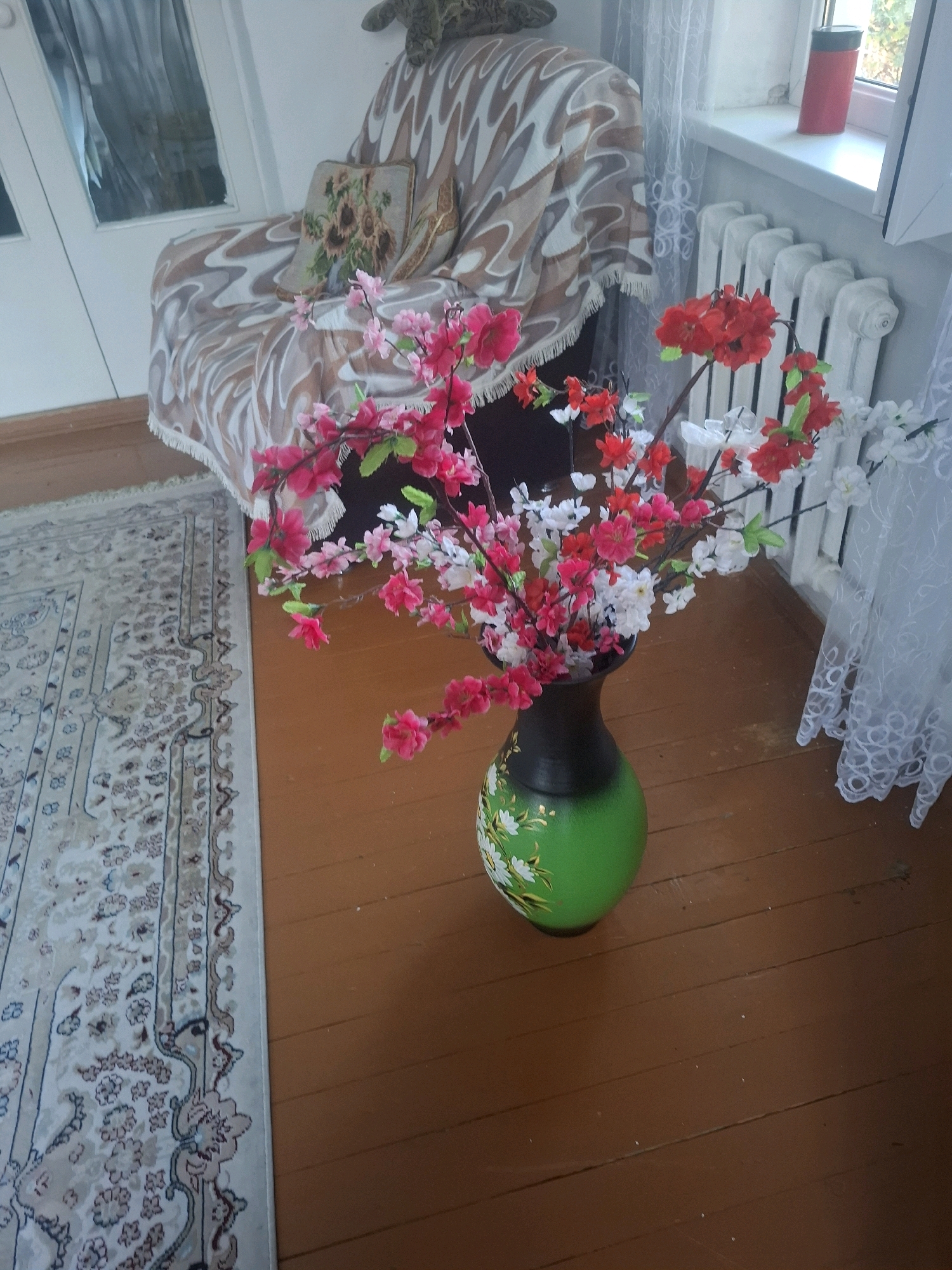 The image depicts an interior scene, likely a residential living room, with no specific geographical indicators placing it in Almaty, Kazakhstan. In the foreground, a vibrant green vase with floral painted details sits on a wooden floor. The vase holds a bouquet of artificial flowers, predominantly in shades of pink and red, with some white blossoms interspersed. To the left of the vase, a portion of a light-colored rug with an intricate pattern is visible. In the background, a sofa is partially visible, draped with a patterned throw blanket. A window with white curtains and a radiator are present to the right. The overall impression is that of a decorated domestic space.