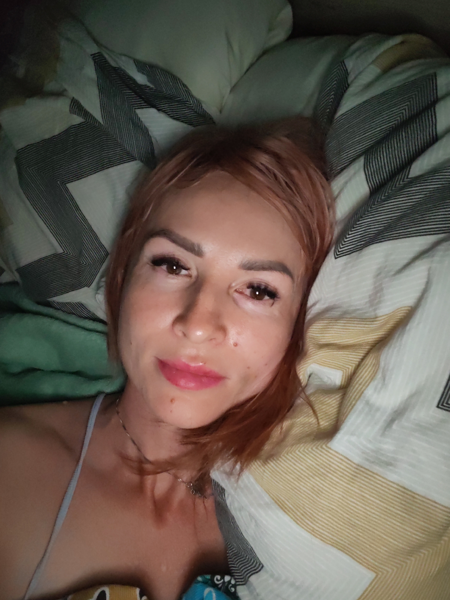 A woman with reddish-blonde hair and makeup is lying in bed. She is wearing a light-colored tank top and a necklace. The bed has pillows with a geometric pattern in black, white, and gold. A green blanket is visible to the left of the woman. The image appears to be a selfie taken in a bedroom. There is no information in the image to suggest this location is in Chaikovsky, Russia.