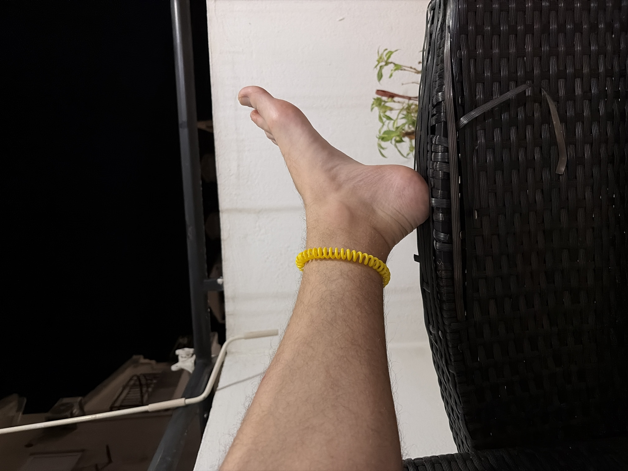 The image shows a leg with a yellow coil bracelet around the ankle, resting on a dark wicker chair. The background includes a white wall with a plant visible and a dark area suggesting nighttime. A white railing and a building are partially visible on the left side of the frame. There is no definitive evidence in the image to confirm that this is Budva, Montenegro.