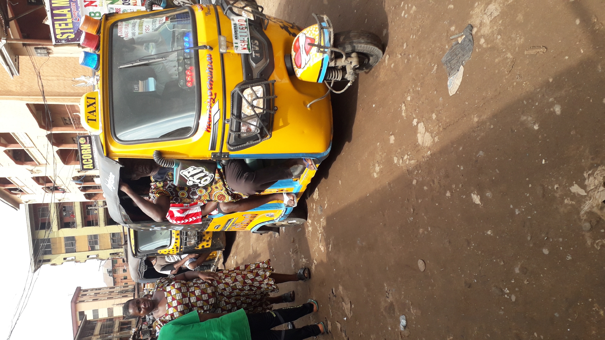 This is a medium shot, eye-level photograph taken in Onitsha, Nigeria. The scene depicts a street with several motorized rickshaws, commonly known as "keke" in Nigeria. In the foreground, a yellow keke is prominently featured. The driver, a man wearing a colorful patterned shirt, is seated inside. The keke has "TAXI" written on its roof and various decals and lights. Two other kekes are visible behind it. Several people are standing or walking around, including a woman in a patterned dress and a person in a green shirt and black pants. The ground is dirt and appears somewhat uneven, with some debris scattered around. Buildings are visible in the background, suggesting an urban environment. The sky is overcast. The image captures a moment of everyday life on a busy street in Onitsha.