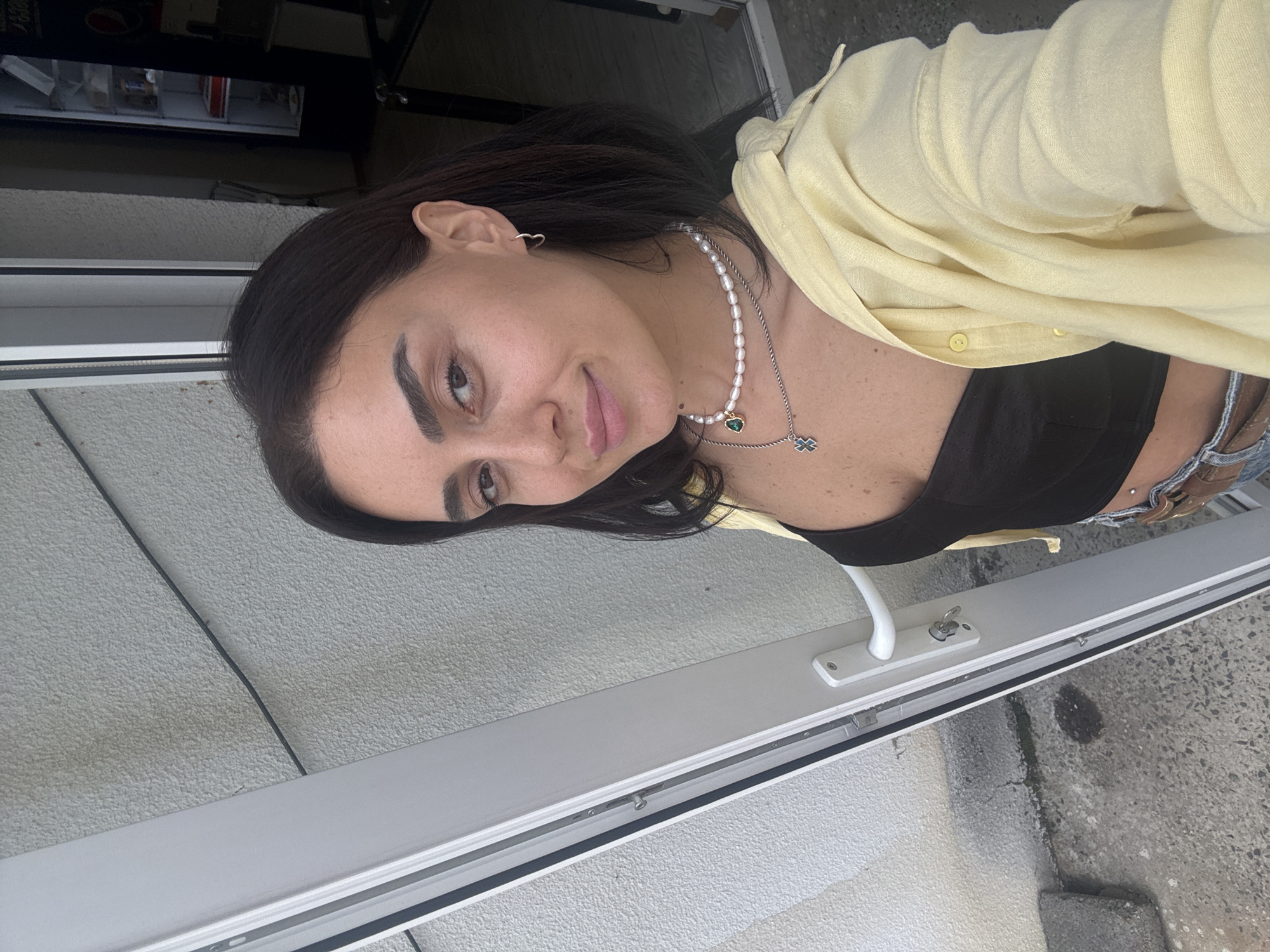 A woman stands in front of a white doorway on a street in Tbilisi, Georgia. She is wearing a black top, a yellow open button-up shirt, and blue jeans. She is also wearing a pearl necklace with a green pendant and a silver chain with a blue cross charm. The woman is looking at the camera and smiling. The background shows a textured white wall, a door handle, and a black vending machine.