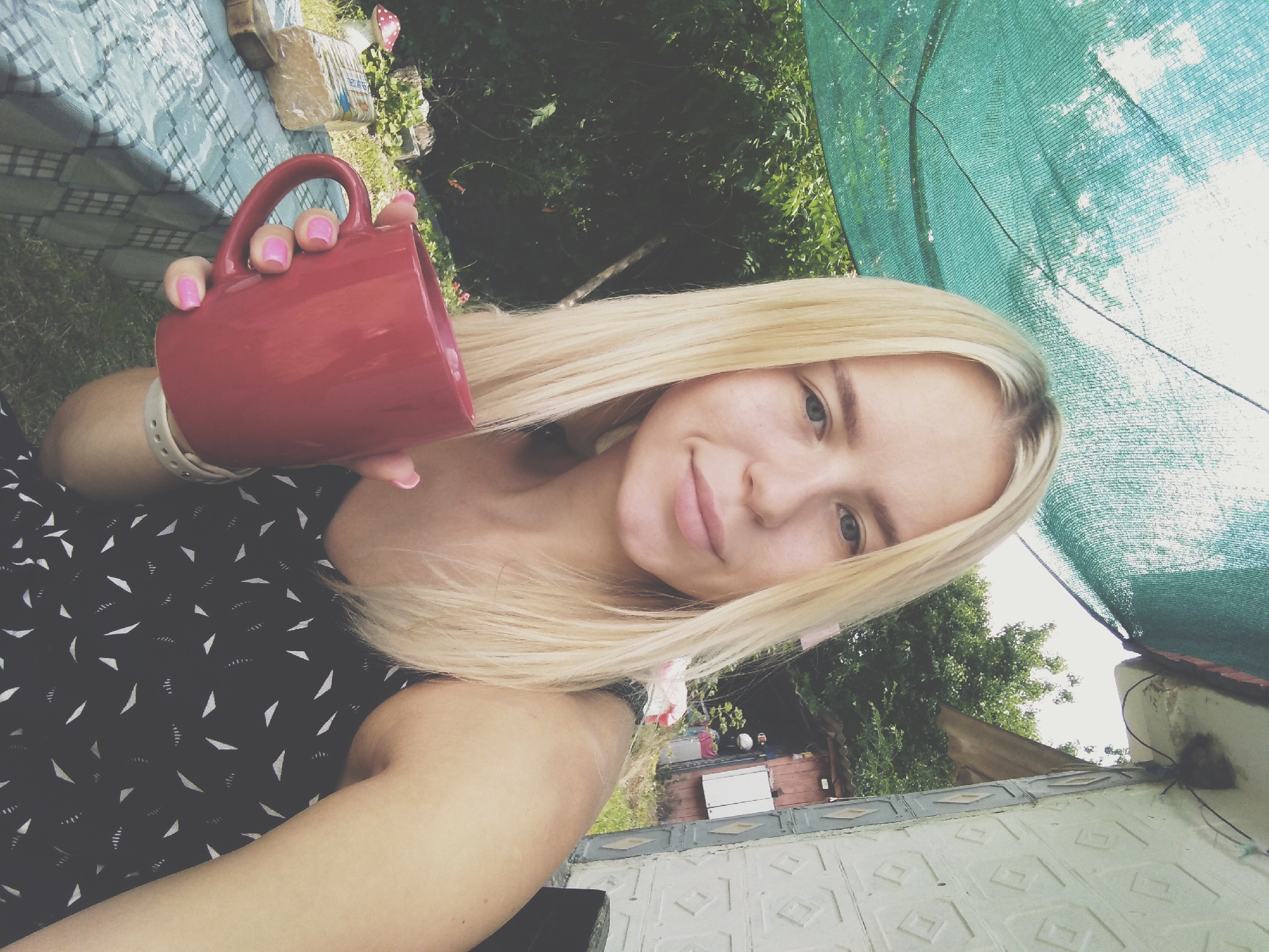 A young woman with blonde hair and blue eyes is taking a selfie outdoors. She is holding a red mug in her right hand. She is wearing a black dress with a white geometric pattern. She has pink nail polish on her fingernails. Behind her, there is a table covered with a blue and white checkered tablecloth, on which are some items, including a package wrapped in plastic and a small mushroom-shaped ornament. A green shade cloth is stretched above the scene, and trees and a building are visible in the background. The location is identified as Radisne, Ukraine.