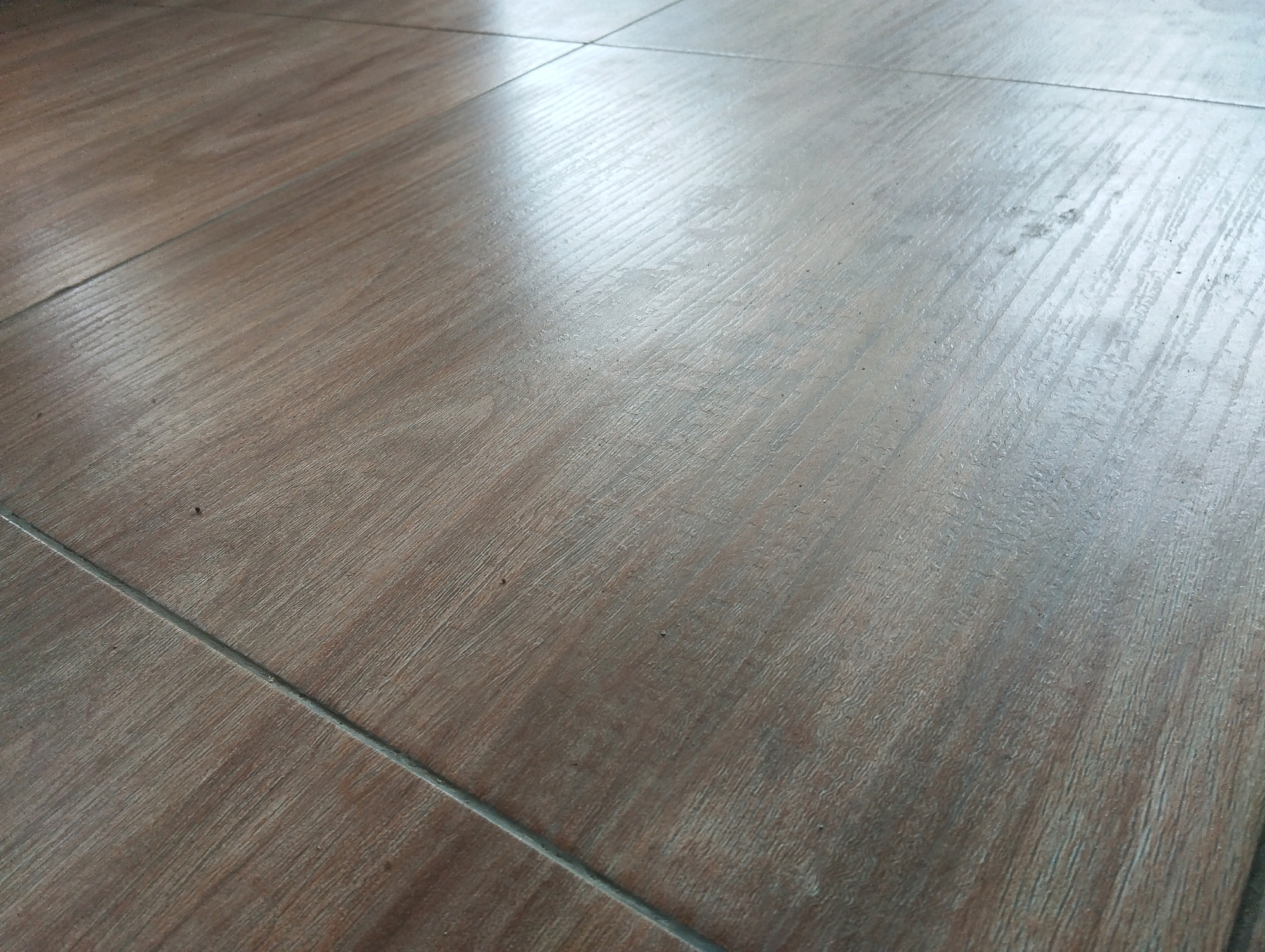 The image shows a close-up, angled view of a tiled floor with a wood-grain pattern. The tiles are laid in a grid, with visible grout lines. The surface is somewhat reflective, with a bright light source creating a glare across the upper right portion of the image. The color of the tiles is a muted brown with variations in tone that mimic natural wood. There are no people or specific objects visible in the frame. The scene is static and focuses on the texture and material of the flooring. This image does not depict any specific location in Jatirejo, Indonesia, as it is a generic view of a floor.