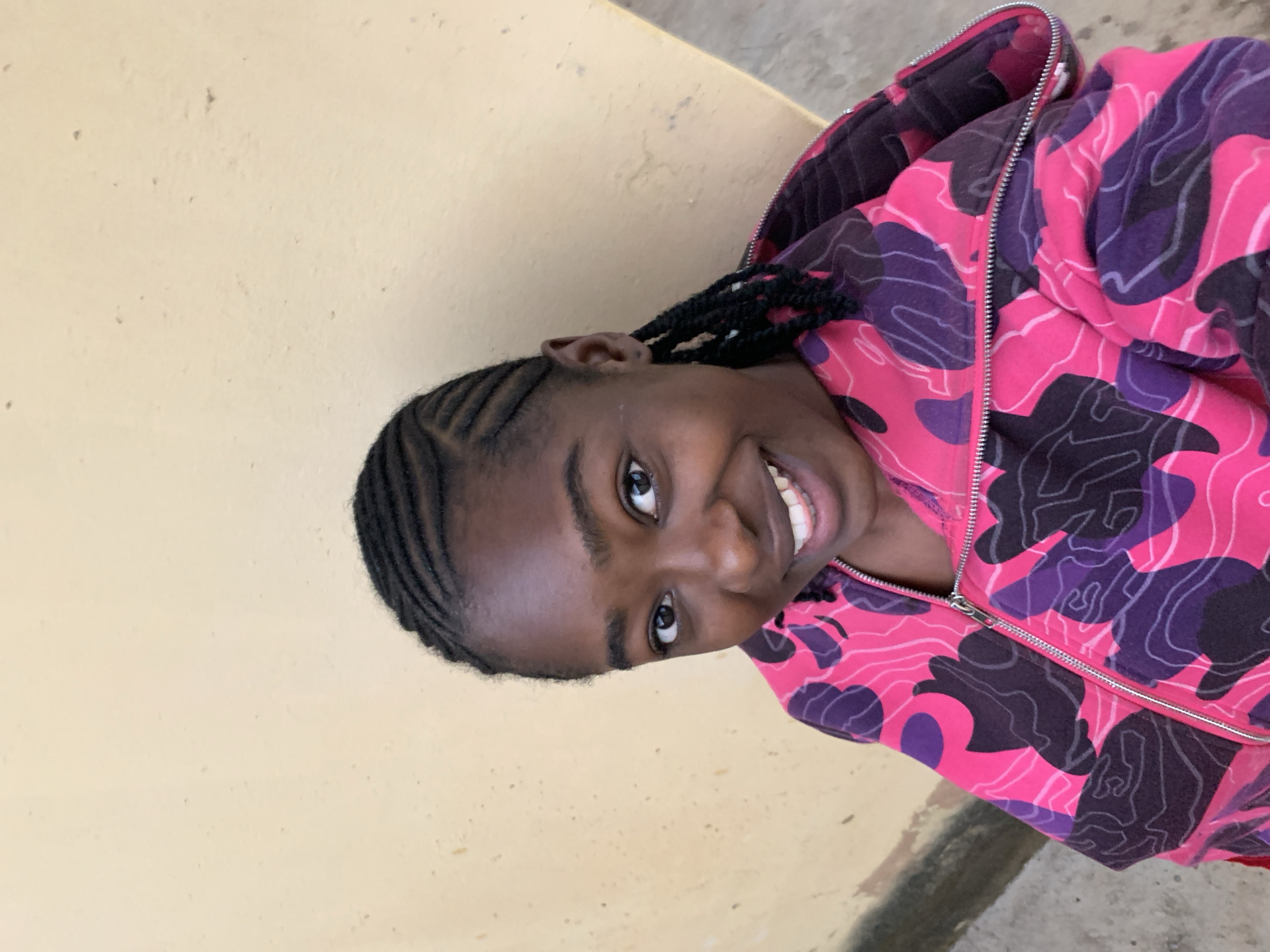 A young person with dark skin and braided hair smiles brightly, looking directly at the viewer in this portrait taken in Rafin Par, Nigeria. Their hair is intricately styled in cornrows leading back from the forehead. They are wearing a hooded jacket featuring a distinctive, vibrant camouflage-like pattern in shades of pink, purple, and black. A silver zipper runs down the front of the jacket, partially open. The person's wide smile reveals white teeth, and their eyes are engaged and cheerful. The background is a plain, light beige or yellowish wall, possibly concrete, with minor imperfections and a slightly darker line at the bottom, suggesting a floor or base. The lighting is soft and even, indicating daytime, but without harsh shadows that would suggest direct sunlight. No text is visible in the image. The overall mood conveyed is one of happiness and approachability.