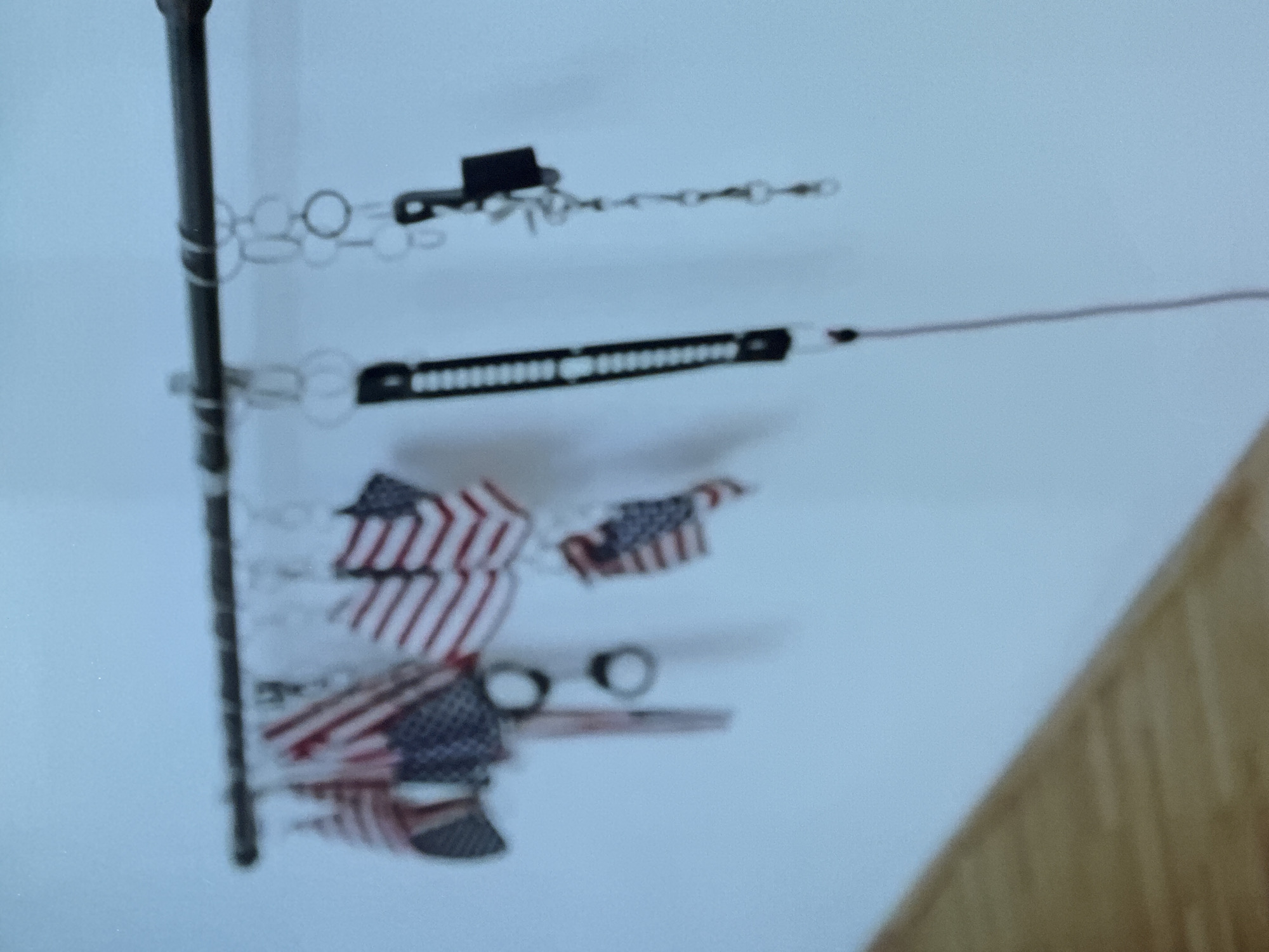 A horizontal, dark-colored metal rod is mounted against a plain, light blue wall. Suspended from this rod are multiple items. On the left, several miniature American flags are displayed, exhibiting the characteristic red and white stripes and a blue canton with white stars. Below some of these flags, a pair of silver-toned metallic handcuffs hangs from a single cuff. Further to the right, another miniature American flag is visible. To the right of the flags, various metallic chain links and rings are hanging. One prominent item is a long, dark, possibly segmented strap or lanyard, which has a thin red cord extending downwards from its end, exiting the bottom of the frame. Another metal chain assembly terminates in a dark, rectangular object with a small hook or clasp. The wall surface is uniform and appears to be indoor. The bottom left quadrant of the image shows a blurred section of a light brown, possibly wooden, surface. No people are visible. The image does not provide any contextual information to establish a specific location within the city of Riga, Latvia.