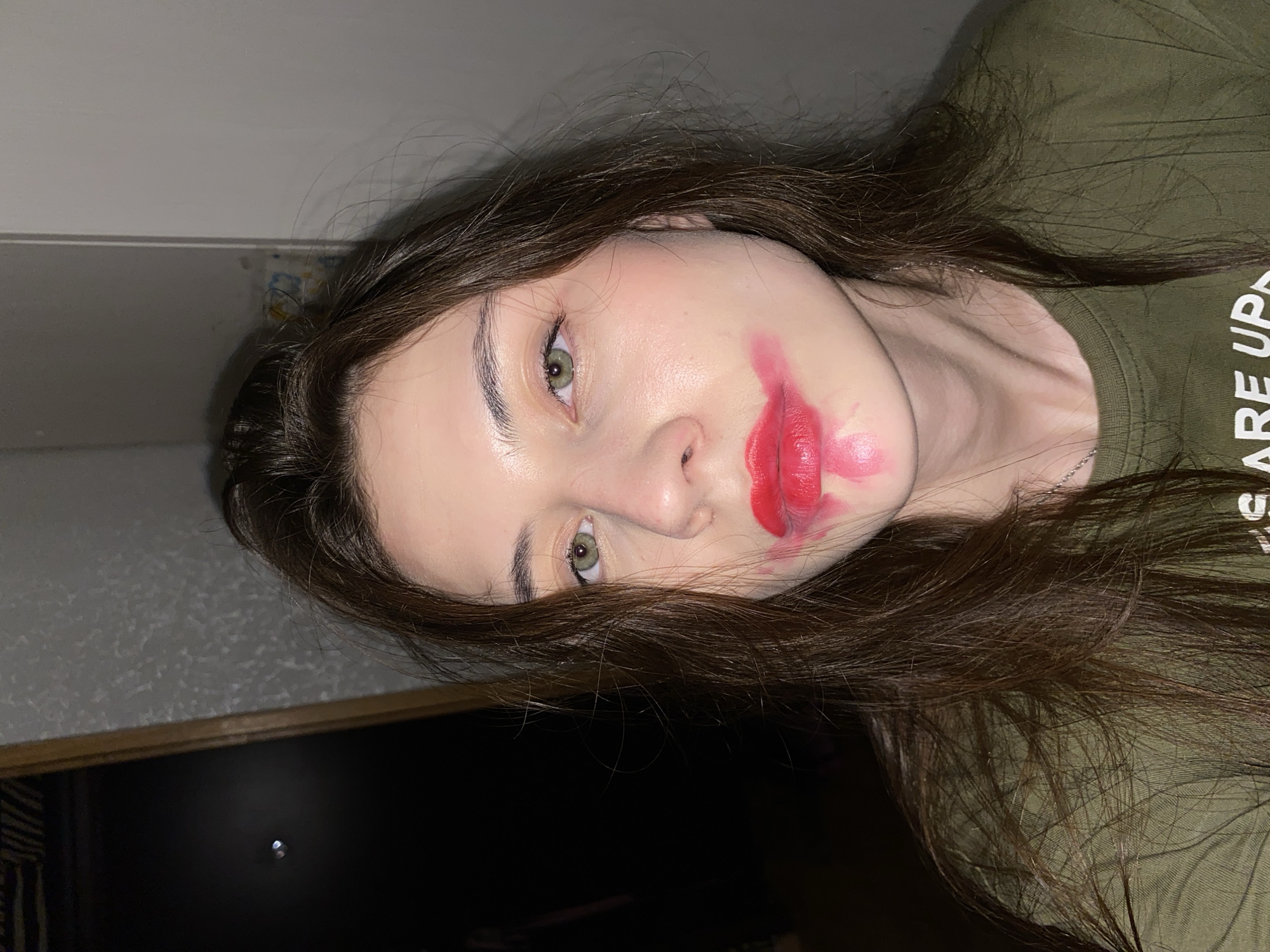 A woman is lying down, facing the camera. Her long brown hair is slightly disheveled, partially obscuring her face. She is wearing an olive green t-shirt. Bright red lipstick is smeared around her mouth and onto her cheek. The background includes a light-colored wall and a dark doorway or opening. The location is not identifiable as being in Votkinsk, Russia based on the image.
