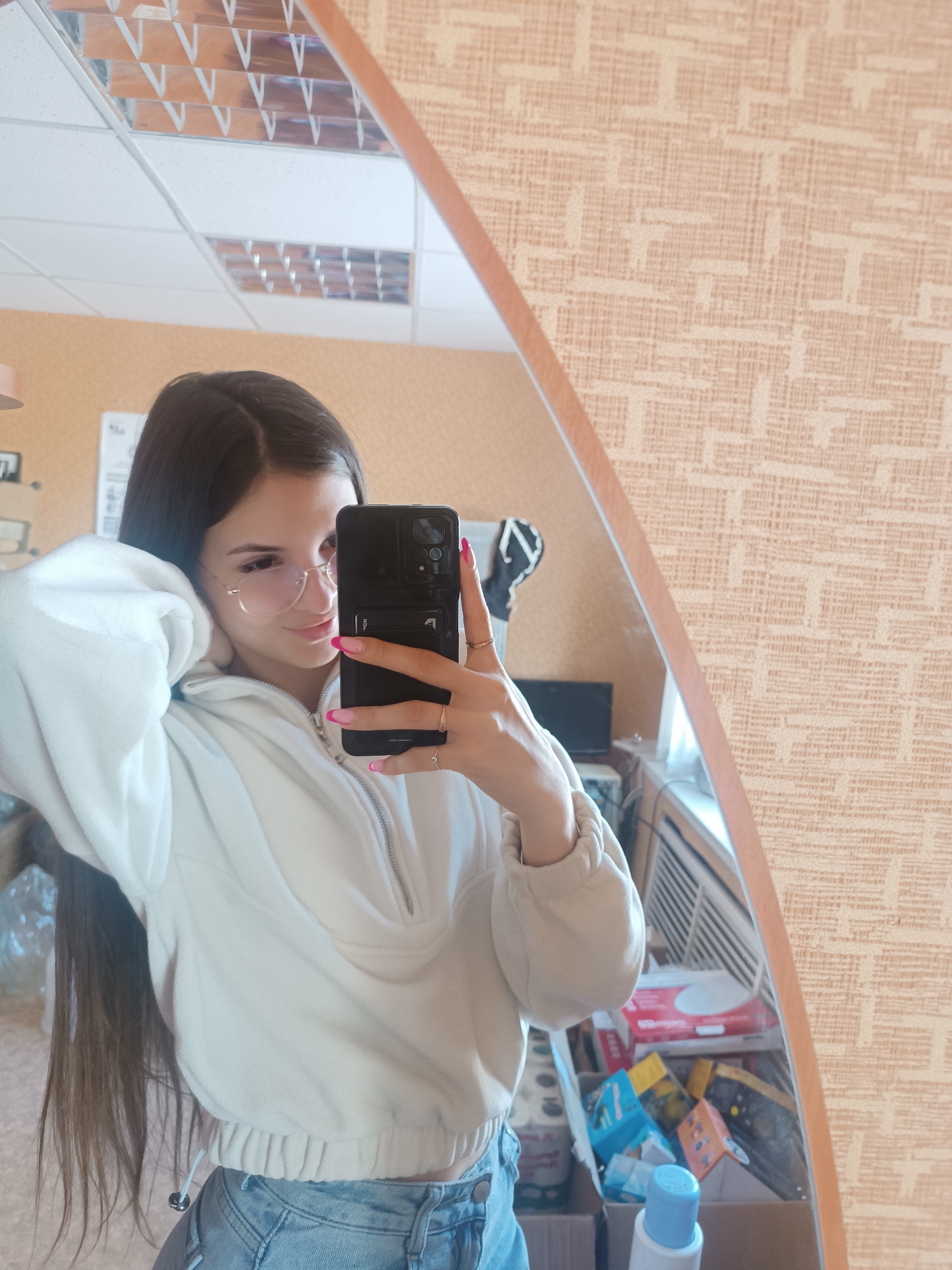 A young woman is taking a selfie in a mirror. She is wearing glasses, a white hooded sweatshirt, and blue jeans. She has long, dark hair. Her nails are painted pink with a white accent. The background includes a textured wallpaper, a ceiling with fluorescent lights, and a shelf with various items, including boxes and bottles. The location appears to be indoors, possibly in a private residence in an urban setting. The city of Unknown, Unknown is not recognizable based on the image.