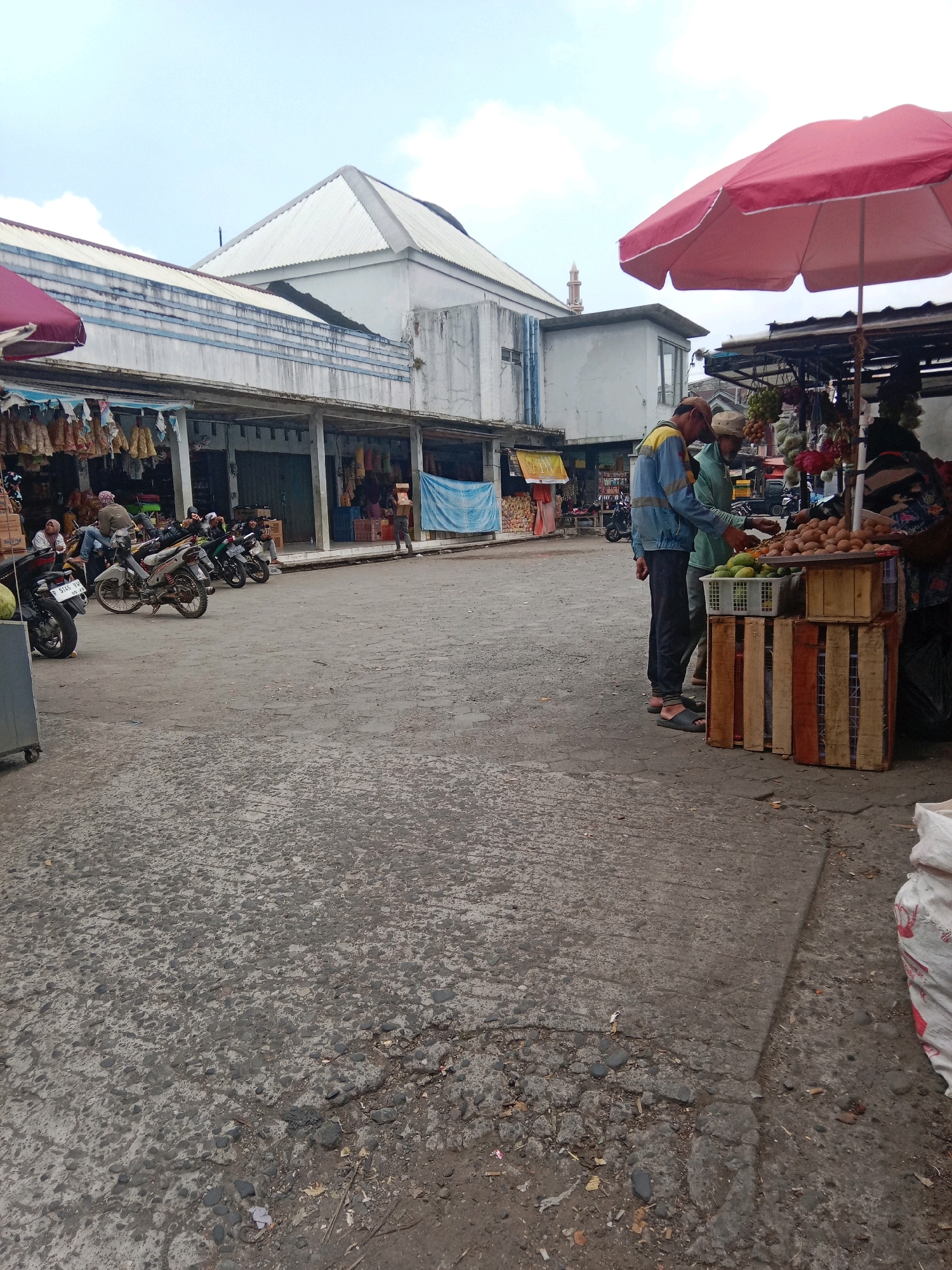 This image captures an outdoor marketplace scene in Batur, Indonesia. In the foreground, the ground is a textured, grey asphalt with visible cracks and scattered debris. To the right, a large white sack is partially visible.
The middle ground features several motorcycles parked on the asphalt. Behind the motorcycles, a row of market stalls lines a building with an open-fronted structure supported by columns. The stalls display various goods, including what appears to be clothing or textiles hanging from the ceiling. Further back, more market structures with awnings are visible.
On the right side of the image, a vendor stand is set up under a large red umbrella. Two individuals, likely customers, are interacting with the vendor, who is partially obscured. The stand displays a variety of produce, including a white basket filled with green fruits, and wooden crates filled with brown, egg-shaped items, possibly potatoes or similar produce.
In the background, a distinctive building with a white, angular roof is prominent. Adjacent to it, a tall, slender structure, resembling a minaret, rises against the sky. The sky itself is overcast with white clouds. The overall environment suggests a bustling local market.