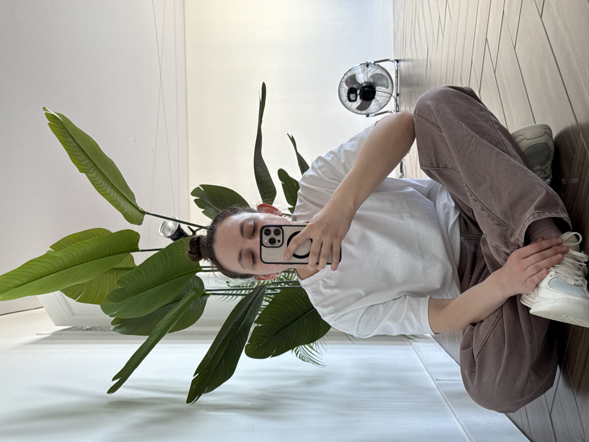 A person is sitting on a floor, taking a selfie with a smartphone. The person is wearing a white t-shirt and brown pants. They are holding a white sneaker in their left hand. To the left of the person is a large plant with green leaves. In the background, there is a white wall and a small fan. The floor appears to be made of white tiles. There is no specific evidence to determine if this image was taken in the city of Chaikovsky, Russia.