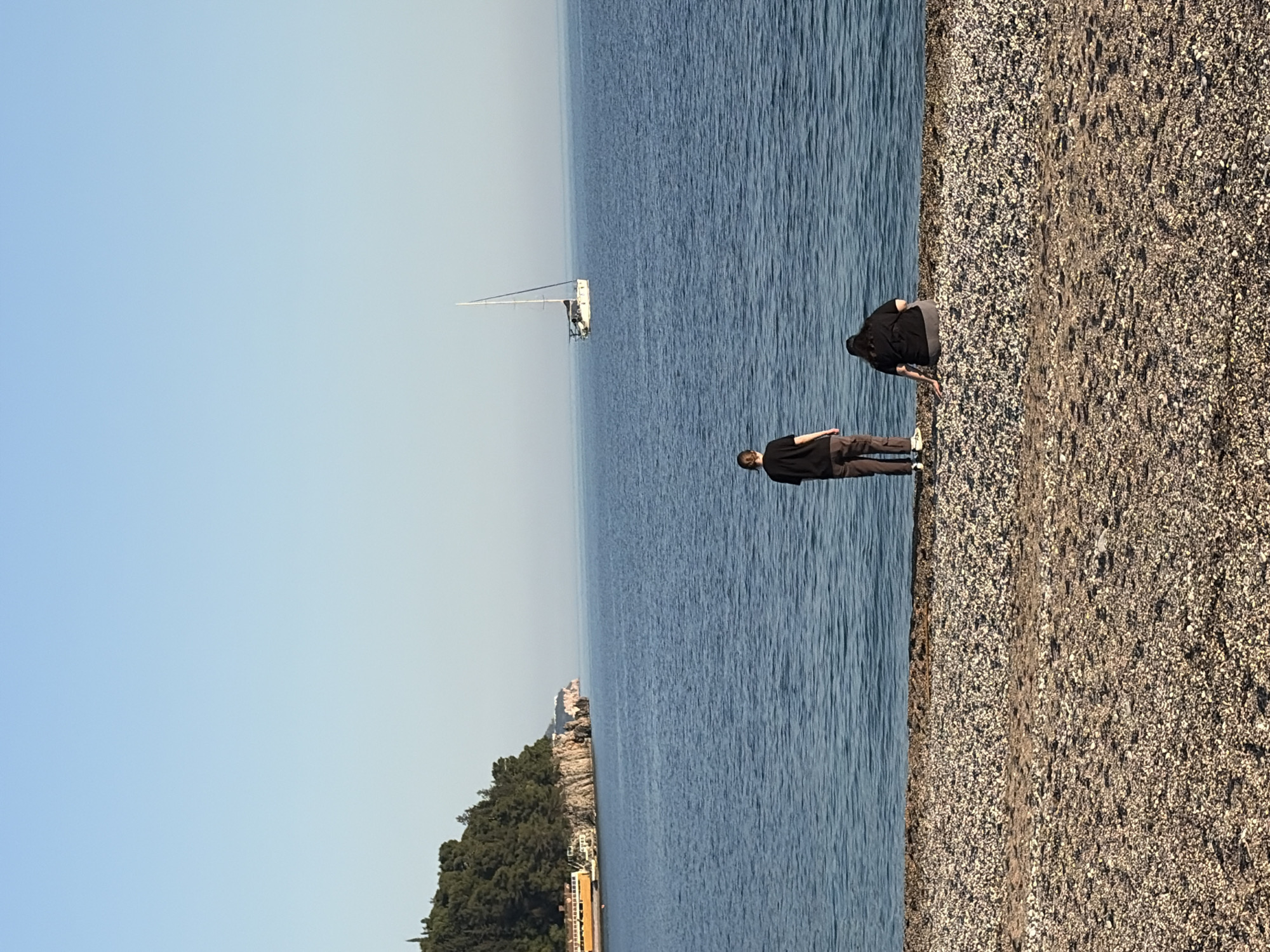 The photograph captures a serene coastal scene, likely taken during daylight hours under a clear sky. The composition is divided into three distinct sections: the sky, the water, and the shoreline. The sky occupies the upper left, transitioning into the vast expanse of the sea, which stretches from the middle towards the right. The shoreline, a pebbly beach, runs along the right edge of the frame. Two individuals are positioned on the shoreline, near the water's edge. One person appears to be standing and looking out at the sea, while the other is bent over, possibly examining something on the beach. A sailboat is visible in the distance on the water. In the lower-left corner, trees and buildings line the land, indicating a possible urban area. The location is not identified.