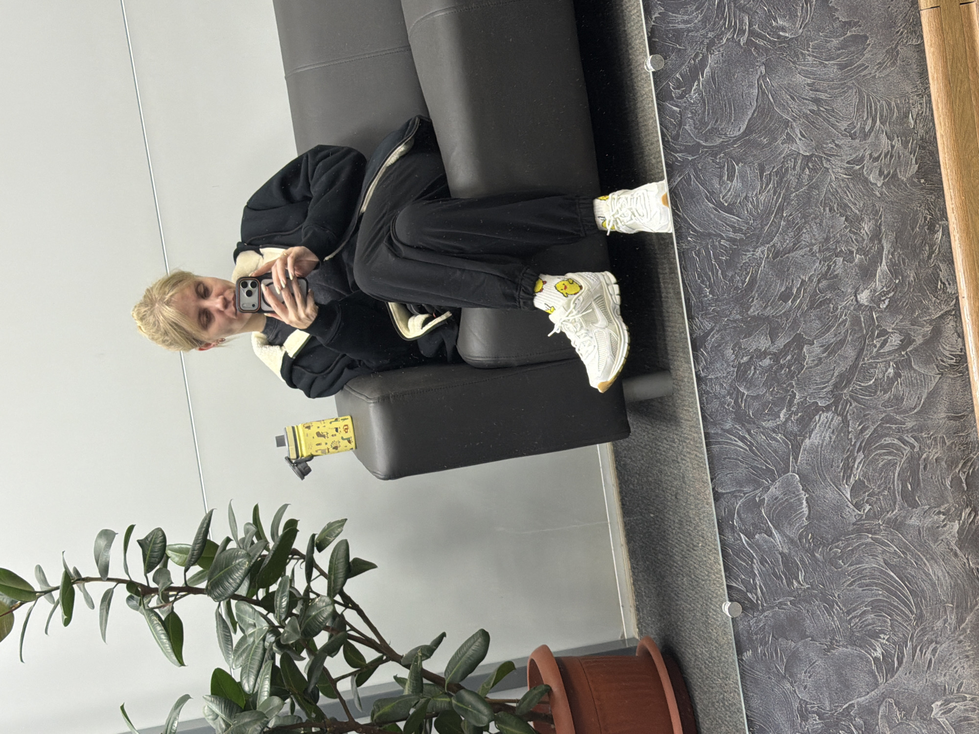 The image shows a person with blonde hair sitting on a black couch, taking a picture with a smartphone in a room that appears to be located in Chaikovsky, Russia. The person is wearing a black jacket, black pants, and white sneakers with cartoon-themed socks. To the left of the couch, there is a green potted plant. In the background, there is a wall with a textured grey design and a wooden frame. The location is likely a public or commercial space based on the decor.