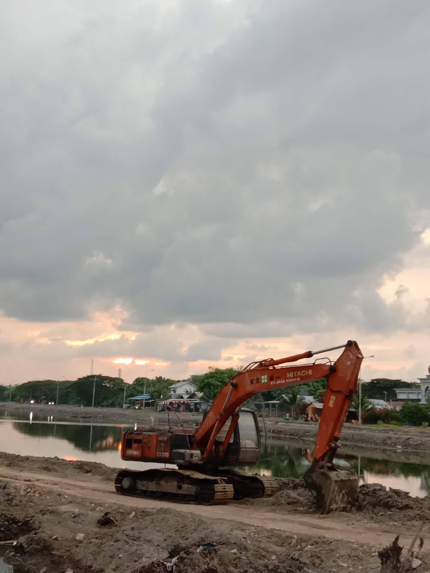 An orange Hitachi EX200 excavator is parked on a dirt path beside a body of water. The excavator has the words "HITACHI" and "CV JASA KUBIKIA" printed on its arm. The sky above is overcast with hints of sunset peeking through the clouds. In the background, there are trees, buildings, and what appears to be a mosque with a dome. The scene suggests a construction or land reclamation project in progress near the coast of Lhokseumawe, Indonesia.