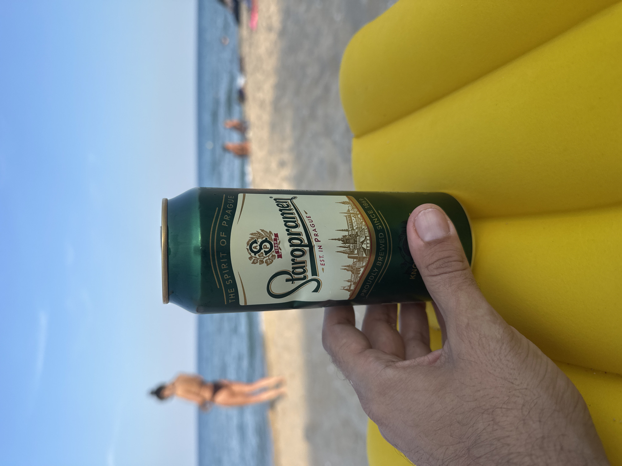 A person's hand holds a can of Staropramen beer on a yellow inflatable object. In the background, people are visible on a sandy beach with the ocean and a clear blue sky. The can is green with a label that says "THE SPIRIT OF PRAGUE" and "STAROPRAMEN EST. IN PRAGUE PROUDLY BREWED SINCE 1869".