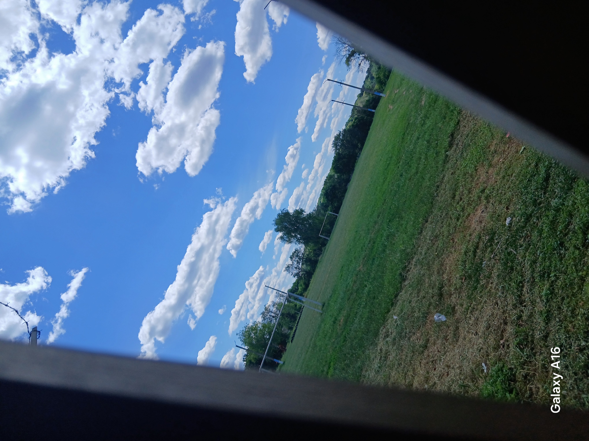 The image depicts an outdoor scene, likely a sports field, situated in Ustyvytsia, Ukraine. The sky above is a vibrant blue, adorned with scattered white cumulus clouds. In the foreground, a grassy field extends into the middle ground, showing areas of both green grass and dried, brown patches. Two soccer goals, constructed with white metal frames, are visible on the field. In the background, a line of trees forms a natural border. The scene is partially framed by dark, diagonal structures, suggesting the viewpoint is from behind a fence or a similar obstruction. The overall impression is of a serene, daytime setting in a rural or semi-rural area.