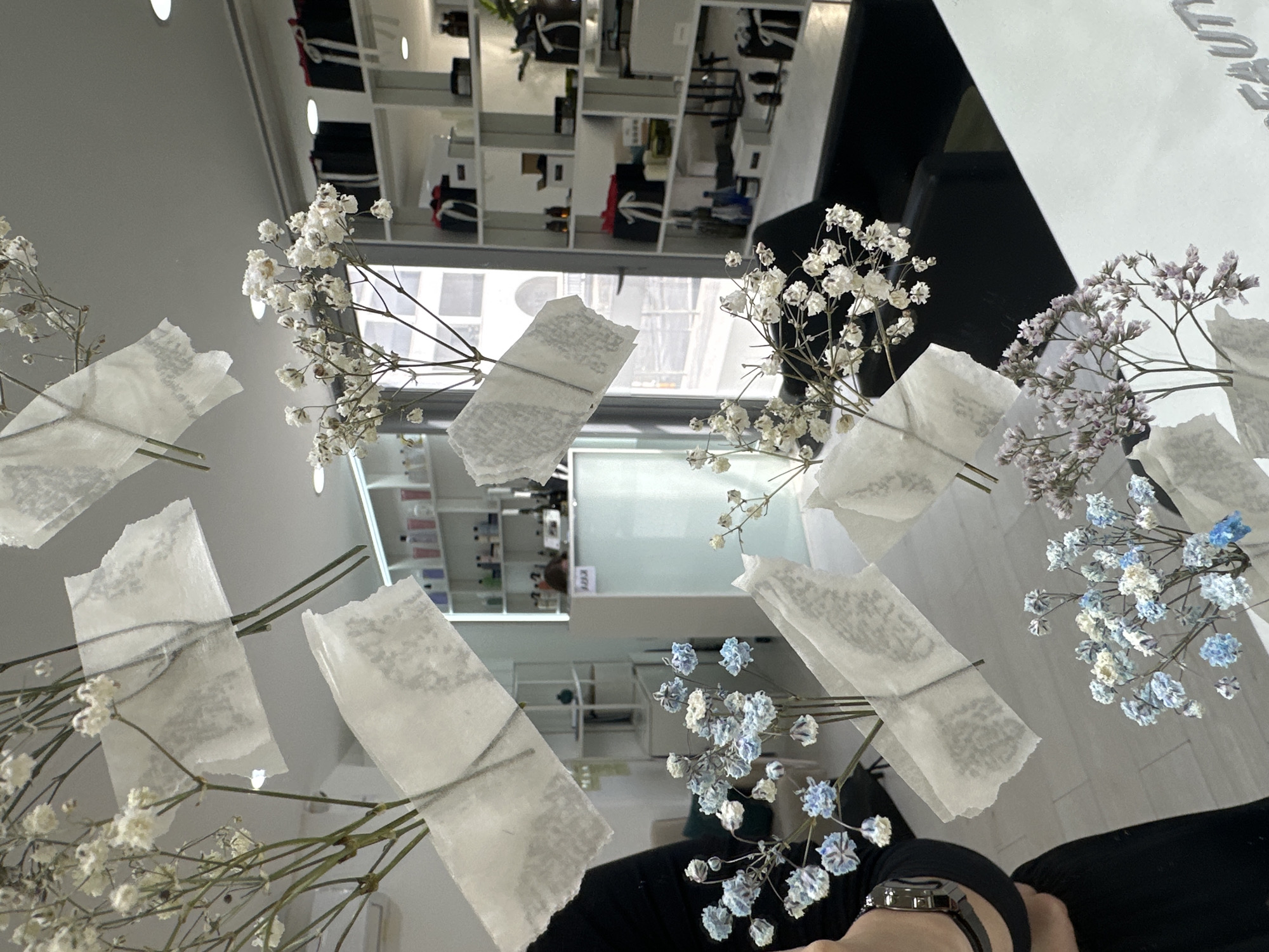 The image depicts a reflective surface, presumably a table or counter, on which are arranged several sprigs of baby's breath flowers secured with small pieces of translucent tape. The flowers are of various colors, including white, blue, and light purple. The surface reflects the interior of a store or salon with shelving units filled with products. The location is possibly Budva, Montenegro, given the surrounding clues. The presence of a person wearing a watch and black clothing suggests a hand or arm is holding the flowers or is nearby. The scene appears to be a staged or styled arrangement for a photograph.