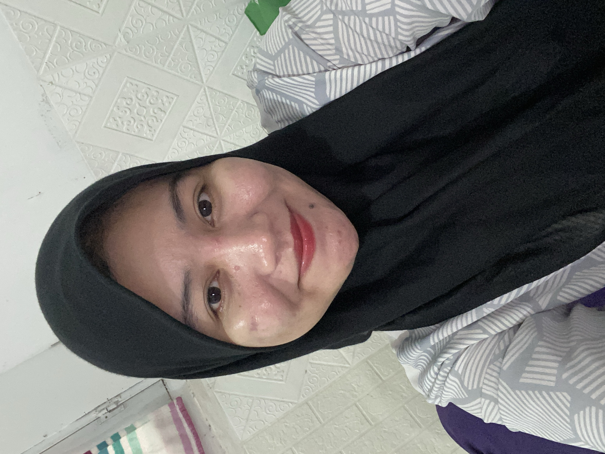 A woman wearing a black hijab and a grey and white patterned shirt is taking a selfie in a room. The background features a white textured wall with geometric patterns and a striped cloth hanging on the left. The woman is smiling and looking at the camera. The lighting suggests an indoor setting, possibly a bedroom or living room. No specific location in Banjarbaru, Indonesia, can be determined from the image alone.