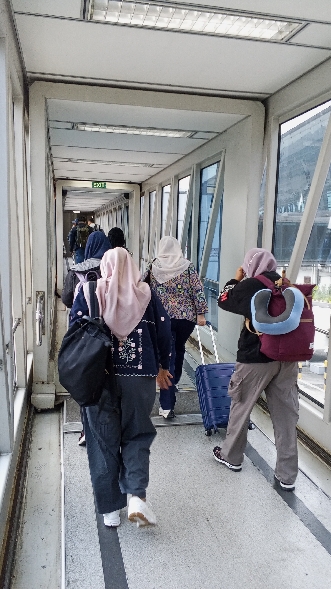 In Rawa Burung, Indonesia, a group of individuals are walking through an airport jet bridge. The jet bridge has large windows on the right side, offering a view of an airport terminal and aircraft. The floor of the jet bridge is grey with black lines, and overhead lighting panels are visible. Several people are walking away from the camera, some pulling suitcases. One person in the foreground is wearing a dark jacket with floral embroidery and a pink headscarf, carrying a black backpack. Another person in the foreground is wearing a dark jacket with a blue neck pillow attached to a burgundy backpack, and grey cargo pants. A blue suitcase is visible on the floor near this person. Further ahead, an "EXIT" sign is illuminated above a doorway. The scene depicts passengers in transit within an airport environment.