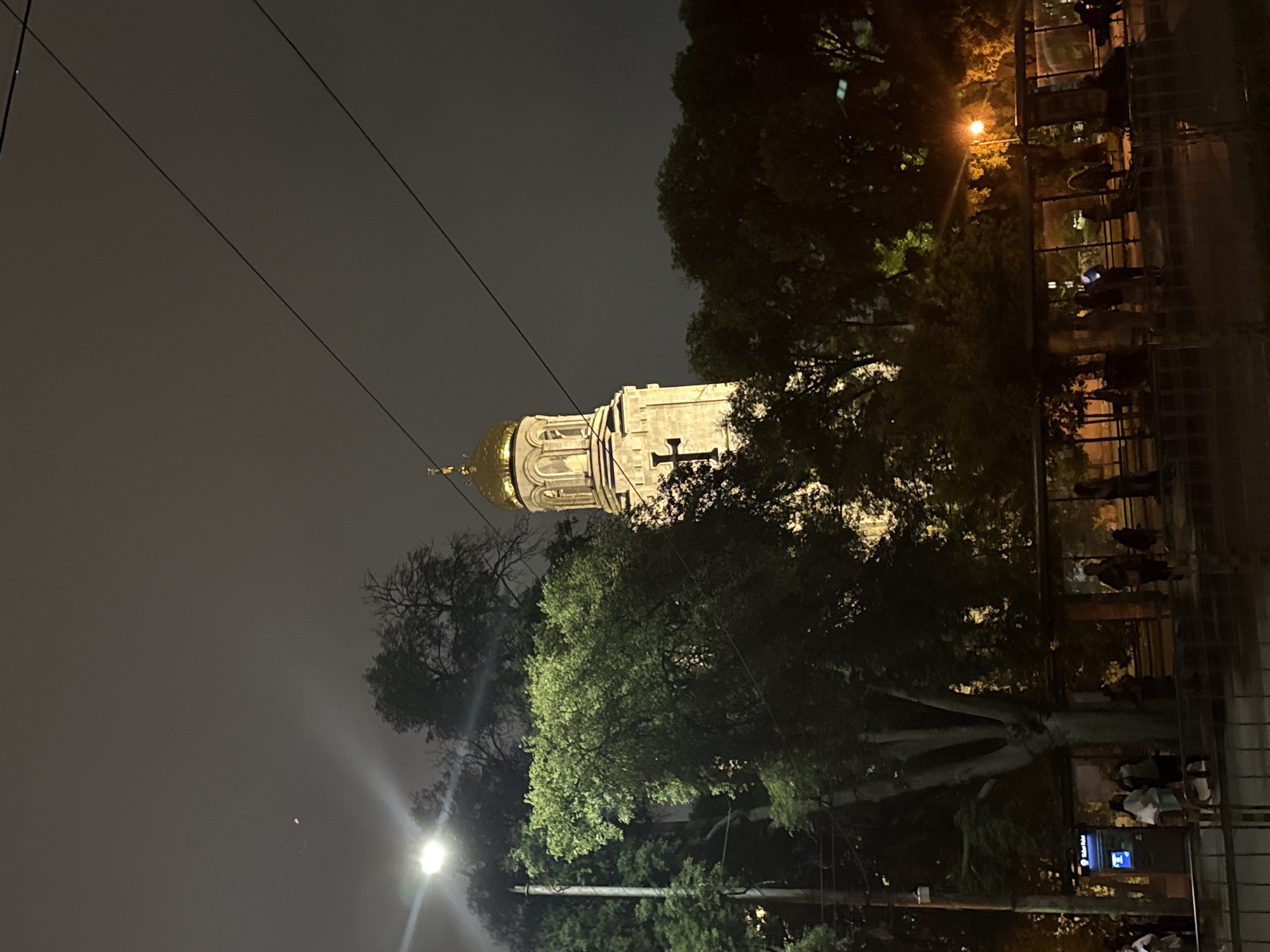 A nighttime urban scene features a prominent, illuminated church tower with a golden dome and a black cross on its side, positioned slightly right of the center. Overcast, dark gray clouds fill the sky, with a bright, distant light source appearing as an orange streak in the upper left. Multiple dark overhead utility lines traverse the upper portion of the frame diagonally.
On the left, a bright white streetlight with significant lens flare illuminates dense green foliage. Numerous trees, some leafy and full, others with sparser branches, are scattered throughout the midground and background, partially obscuring the base of the church tower.
In the lower half of the image, a public transport stop or shelter extends horizontally, populated by approximately 10-15 individuals. They are mostly standing or walking, appearing to wait. A "Ticket Point" machine with a blue illuminated screen is visible on the far left of the shelter area. Another orange-hued streetlight on the far right casts light on additional trees and people at the bus stop. A dark metal railing runs along the bottom edge of the image. The scene is located in an unknown city, unknown country.