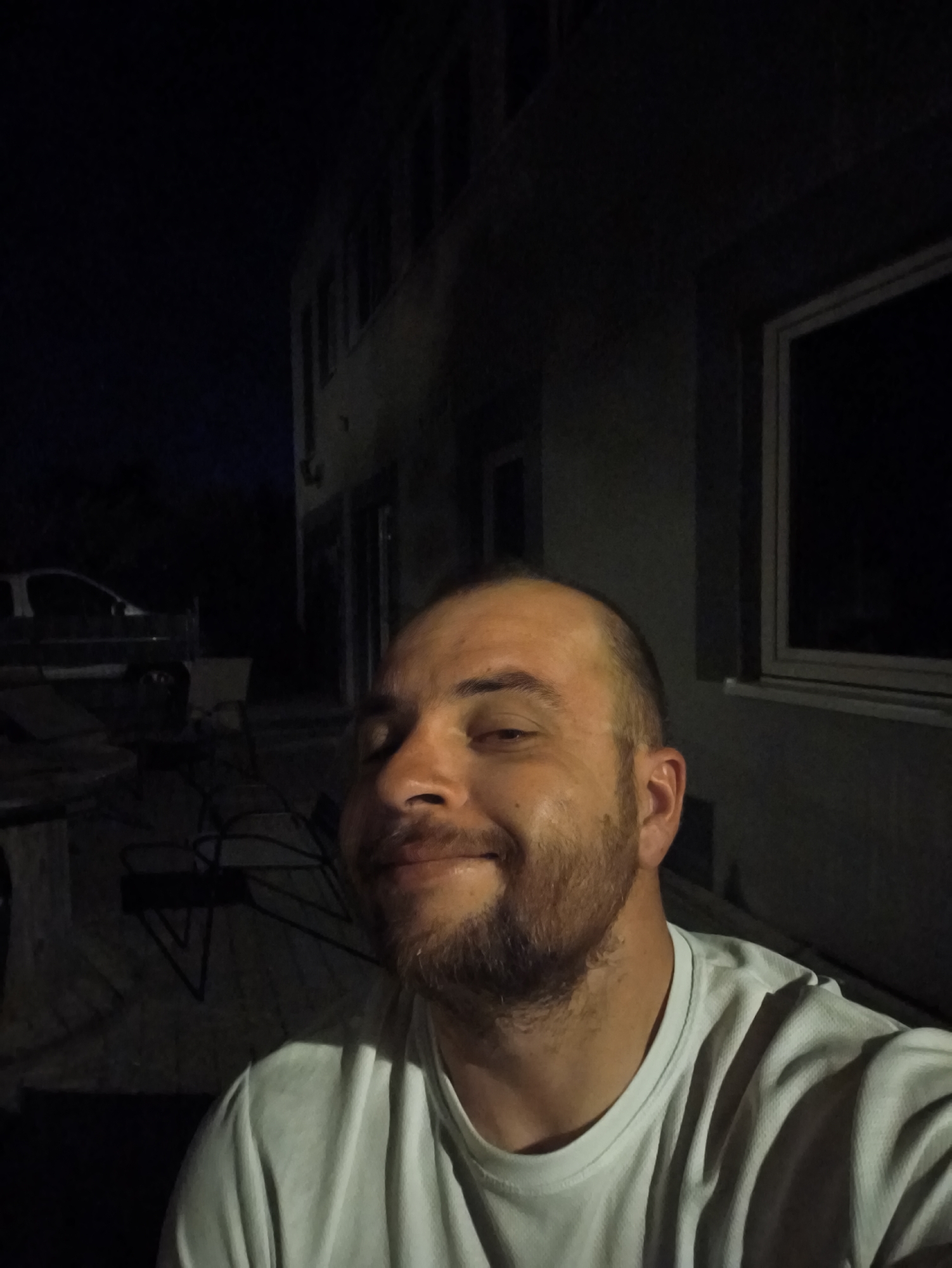At night, a man with a beard and a white t-shirt is standing outside a building with windows. A car is parked in the background. The location is Unknown, Unknown.