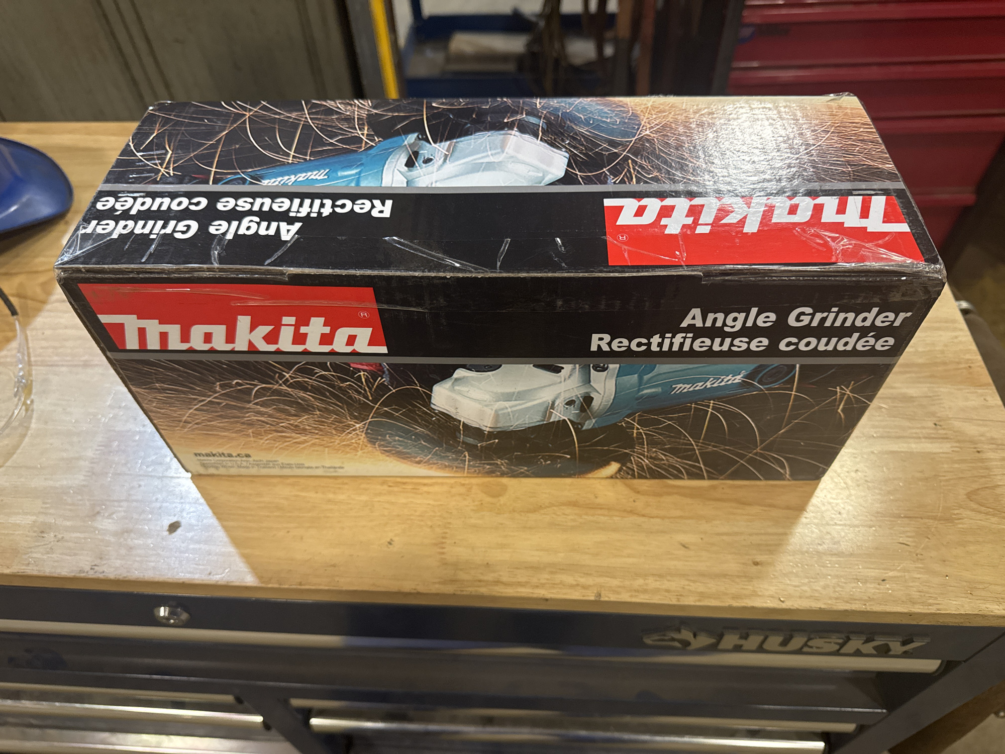 The image shows the top and side of a cardboard box for a Makita angle grinder, placed on a wooden workbench. The box is primarily black with a graphic of a grinder and sparks. Text on the box reads "Makita," "Angle Grinder," and "Rectifieuse coudée." The box is sealed with clear tape. To the left of the box, a blue object and a glass or plastic container are partially visible. The background shows a metal cabinet or shelving unit and a blurred industrial setting. The image's location in Calgary, Canada cannot be determined from the visual information provided.