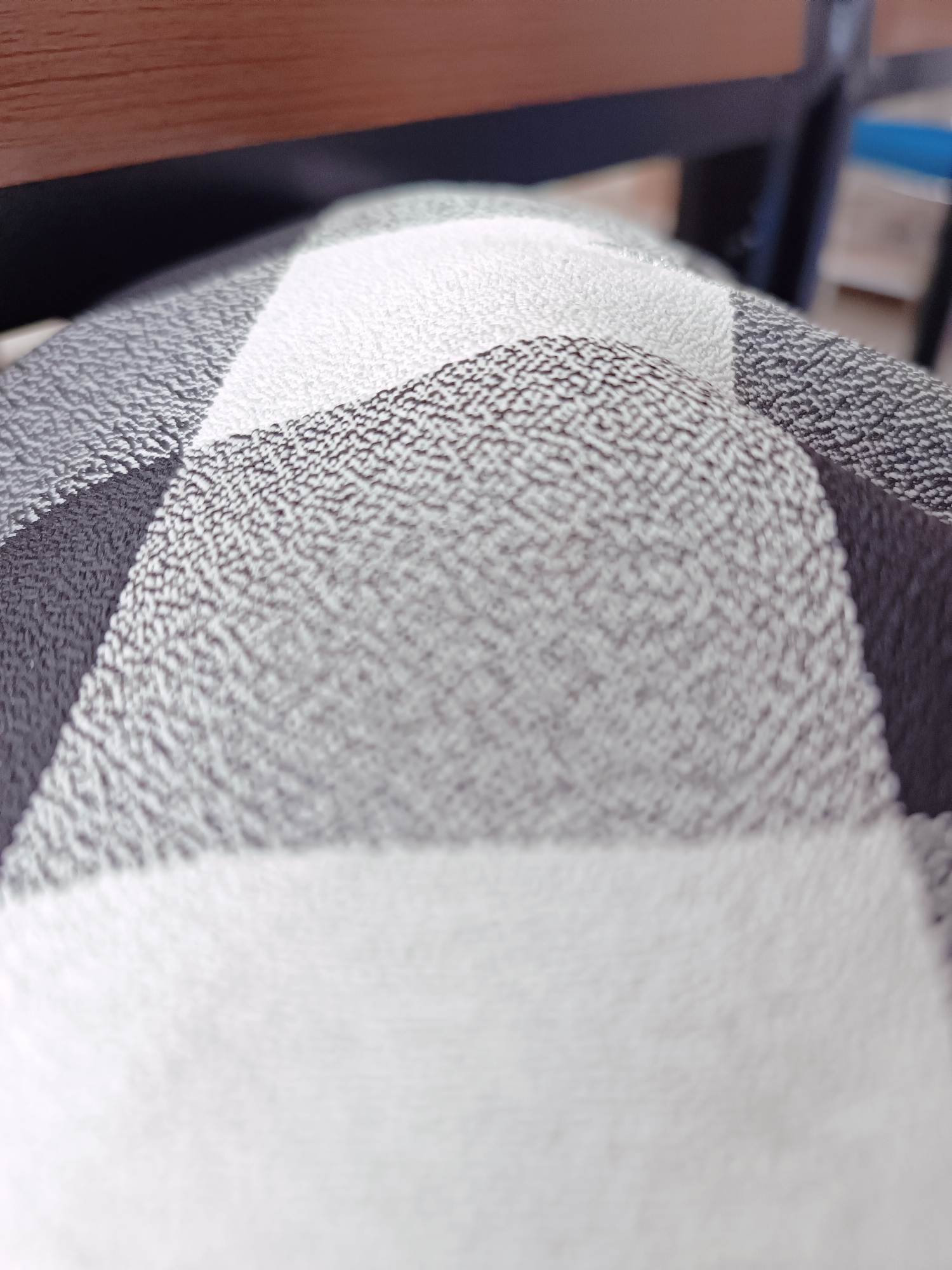 A close-up, low-angle shot captures a textured fabric with a black and white checkered pattern. The fabric is slightly creased, creating subtle shadows and highlights that emphasize its woven surface. In the upper portion of the image, a dark, blurry background suggests the presence of furniture, possibly made of wood, given the visible brown color. The overall scene is abstract and focused on the tactile quality of the material, with no discernible human presence or activity. The specific location in Tabanan, Indonesia, cannot be determined from this image alone.