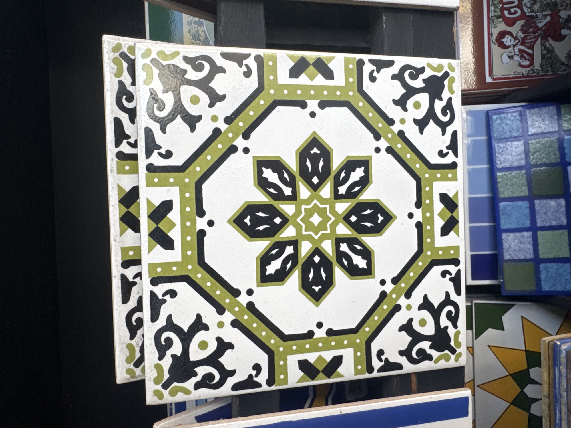 The image shows a close-up view of two decorative tiles stacked on top of each other, with a focus on the top tile. The top tile features an intricate octagonal design with a white background. The pattern consists of a central eight-pointed star composed of black and lime-green shapes, surrounded by a lime-green border with small white dots. The corners of the tile are decorated with black, curvilinear, vine-like motifs. The tile is placed on a dark, blurred background, possibly a shelf or display. The bottom tile is partially visible and appears to have a similar but less distinct pattern. The overall impression is of a decorative object, likely part of a collection or display of tiles. There are no people or actions depicted in the image, and the environment is non-descript and focused on the tiles. It's not possible to determine the exact location in Niterói, Brazil from the image itself.