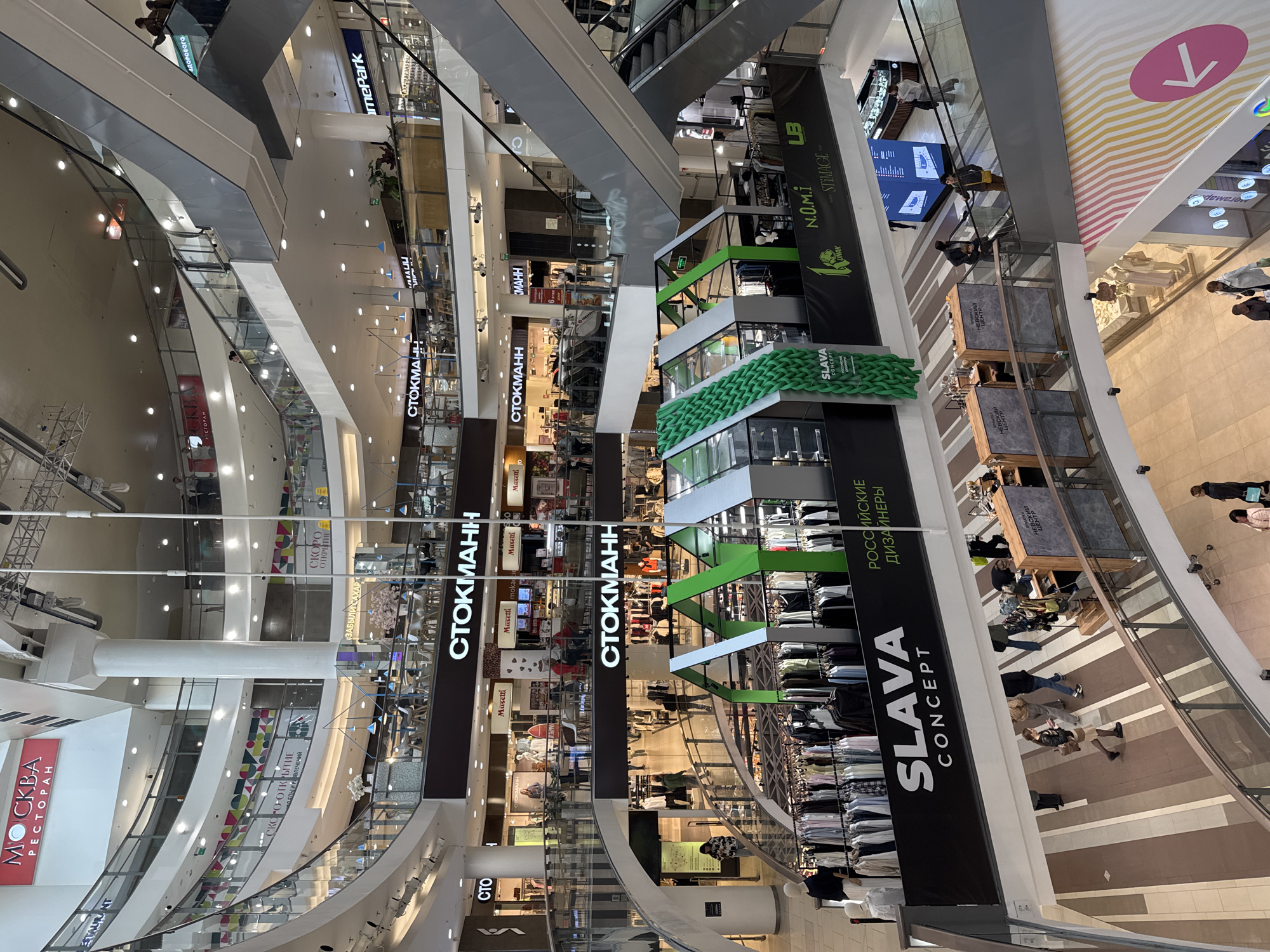 The image depicts the interior of a multi-level shopping mall in Saint Petersburg, Russia. Several stores with signs are visible, including "СТОКМАНН" (Stockmann), "SLAVA CONCEPT," "N.O.M.i. STIMAGE," "HomePark," "dewele," and "LB," indicating various retail and food establishments. "МОСКВА РЕСТОРАН" (Moscow Restaurant) is also visible. Escalators and glass railings are present, facilitating vertical movement within the mall. People are seen walking and browsing within the mall. The environment is brightly lit with overhead lighting. Green decor elements can be seen throughout the mall. The floor has a striped design in tan and brown tones.