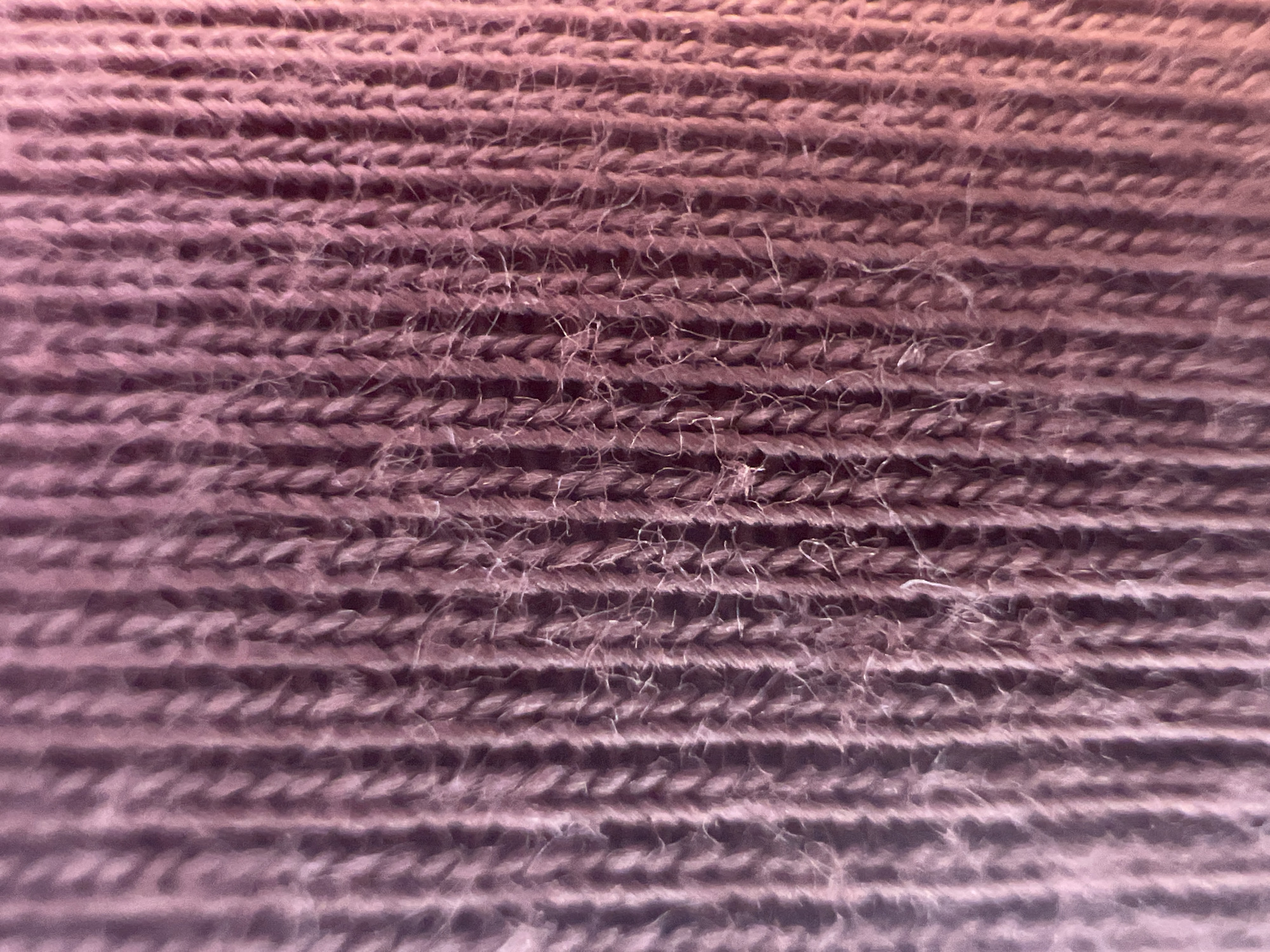 The image is a close-up, macro photograph of a knitted fabric. The fabric appears to be made of fine yarn, possibly wool or a blend, in a muted rose or dusty purple color. The knitting pattern is a simple rib knit, with vertical columns of stitches creating a ribbed texture. Some loose fibers are visible, giving the fabric a slightly fuzzy or worn appearance. The lighting suggests a soft, diffused light source, with subtle gradients of light and shadow highlighting the texture of the knit. There are no people, objects, or environmental elements visible in the image, other than the fabric itself. The image is a detail shot focused entirely on the texture and color of the textile.