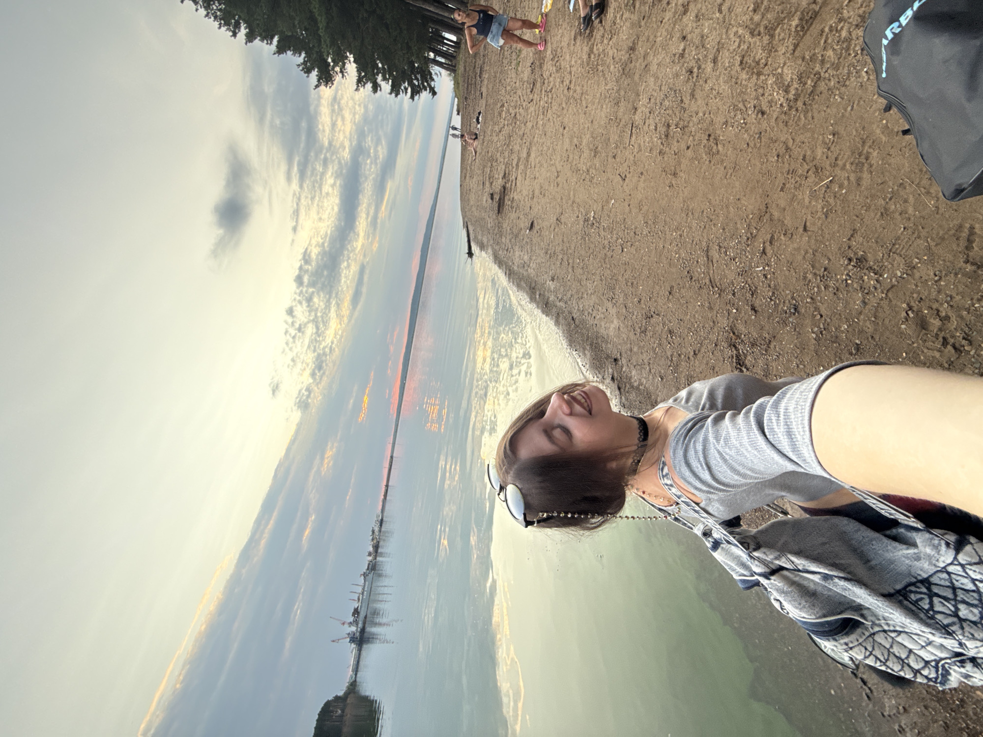 A selfie of a young woman with brown hair pulled back and sunglasses on her head, standing on a sandy beach by the water. She is wearing a grey, off-the-shoulder shirt and a dark choker necklace. A patterned backpack is slung over her left shoulder. In the background, the water stretches to the horizon where a industrial port with cranes and ships is visible. The sky is partly cloudy with a sunset glow. Further down the beach, a few other people are visible, including a woman in a blue top and denim skirt. The environment appears to be a lake or a large body of water with a sandy shore and a line of trees in the distance.