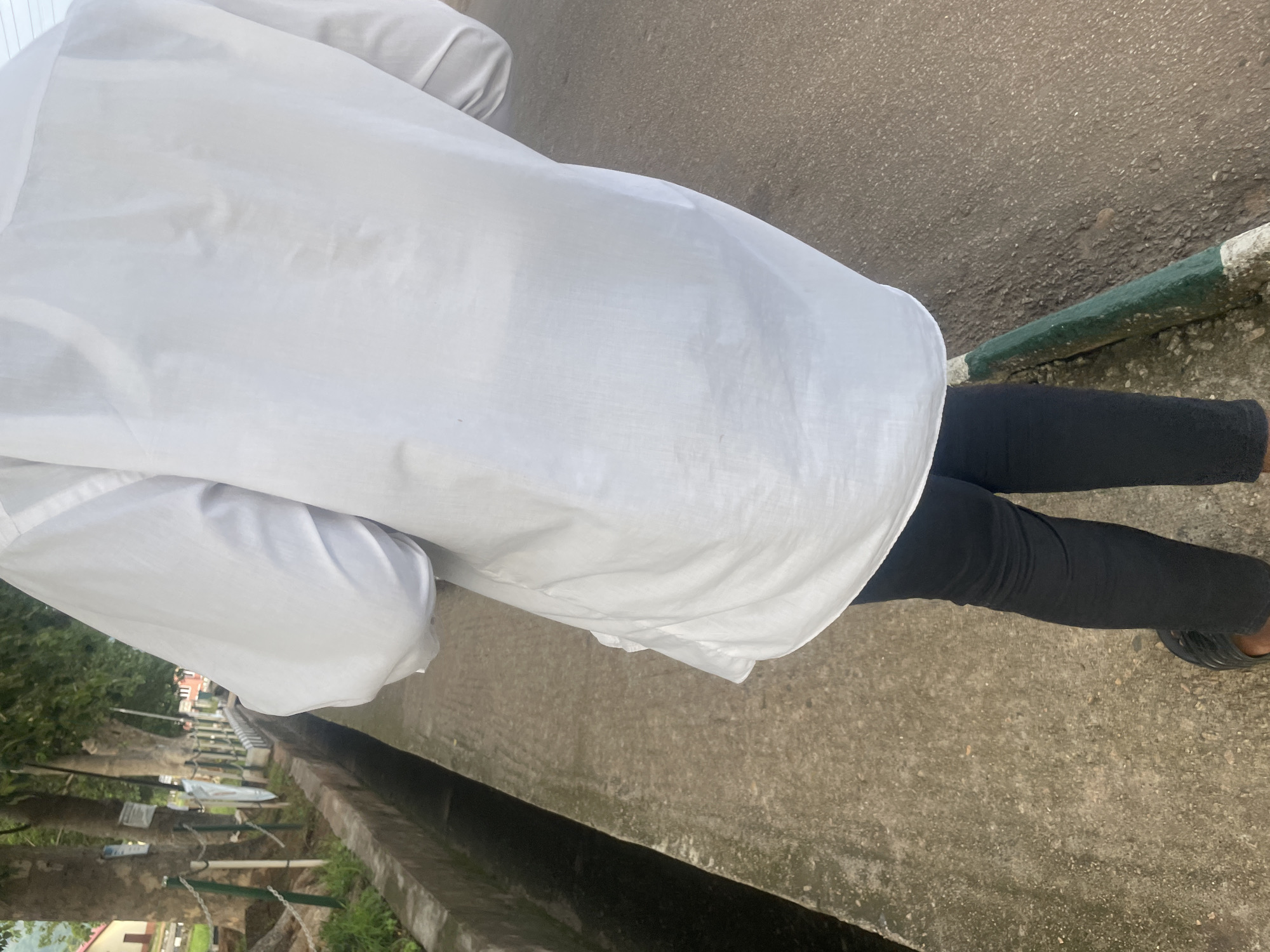 The image shows a person from behind, wearing a white shirt and black pants. They are walking on a concrete path next to a drainage ditch. Trees line the path on the left side, with a building visible in the background. The person is wearing dark sandals. The scene appears to be in an outdoor urban environment.