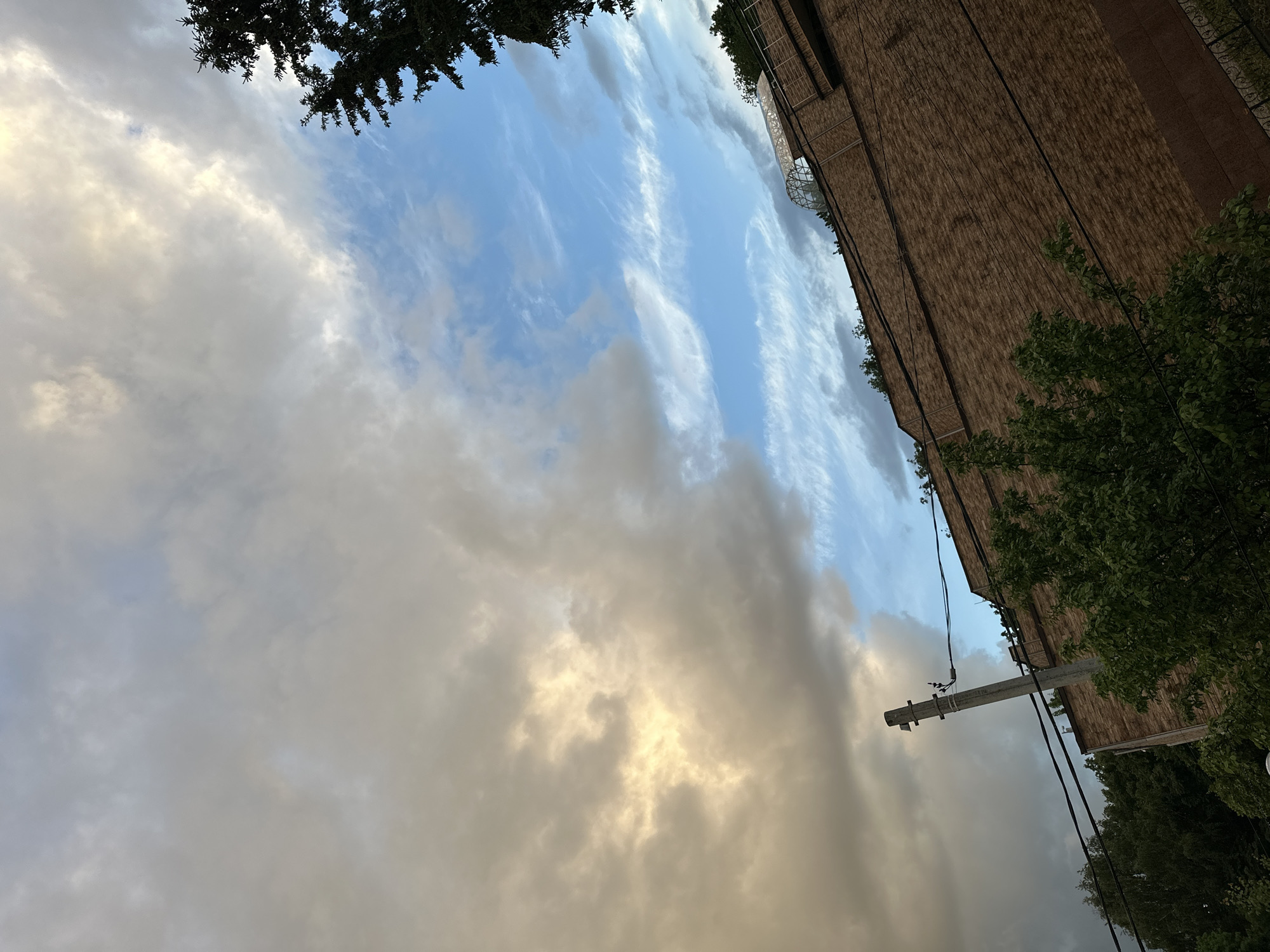 The image shows an upward view in an urban setting, featuring a combination of natural and man-made elements. In the sky, there are two types of clouds: one consists of thin, striated clouds resembling cirrus, and the other is a dense, gray cloud formation. Partial views of trees are visible in the upper-left corner, and green foliage appears on the right side, partially obscuring a building. The building has a brick facade with visible windows and a metal railing. A utility pole with attached wires is positioned against the backdrop of the sky and the building. The overall impression is a dynamic composition of natural and urban features. The image lacks sufficient detail to identify a specific location in Nizhny Novgorod, Russia. There are no people visible in the image.