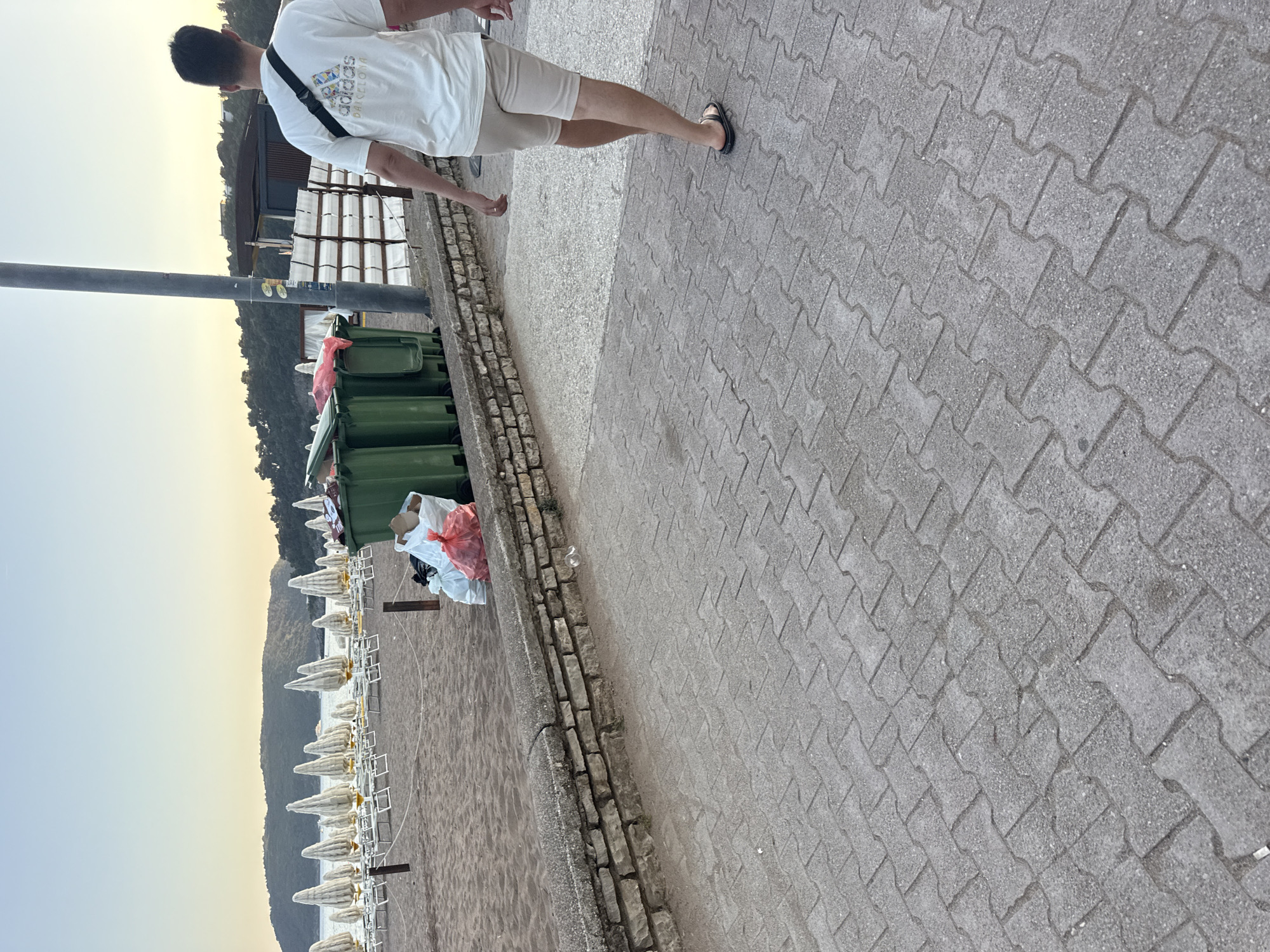 In Boreti, Montenegro, a man walks away from the camera on a paved walkway that borders a sandy beach. The beach has rows of closed white umbrellas and lounge chairs. To the left of the man are several green trash cans overflowing with garbage bags. The man is wearing a white t-shirt and light-colored shorts. In the background, hills covered in trees rise up, and the sky is a pale blue.